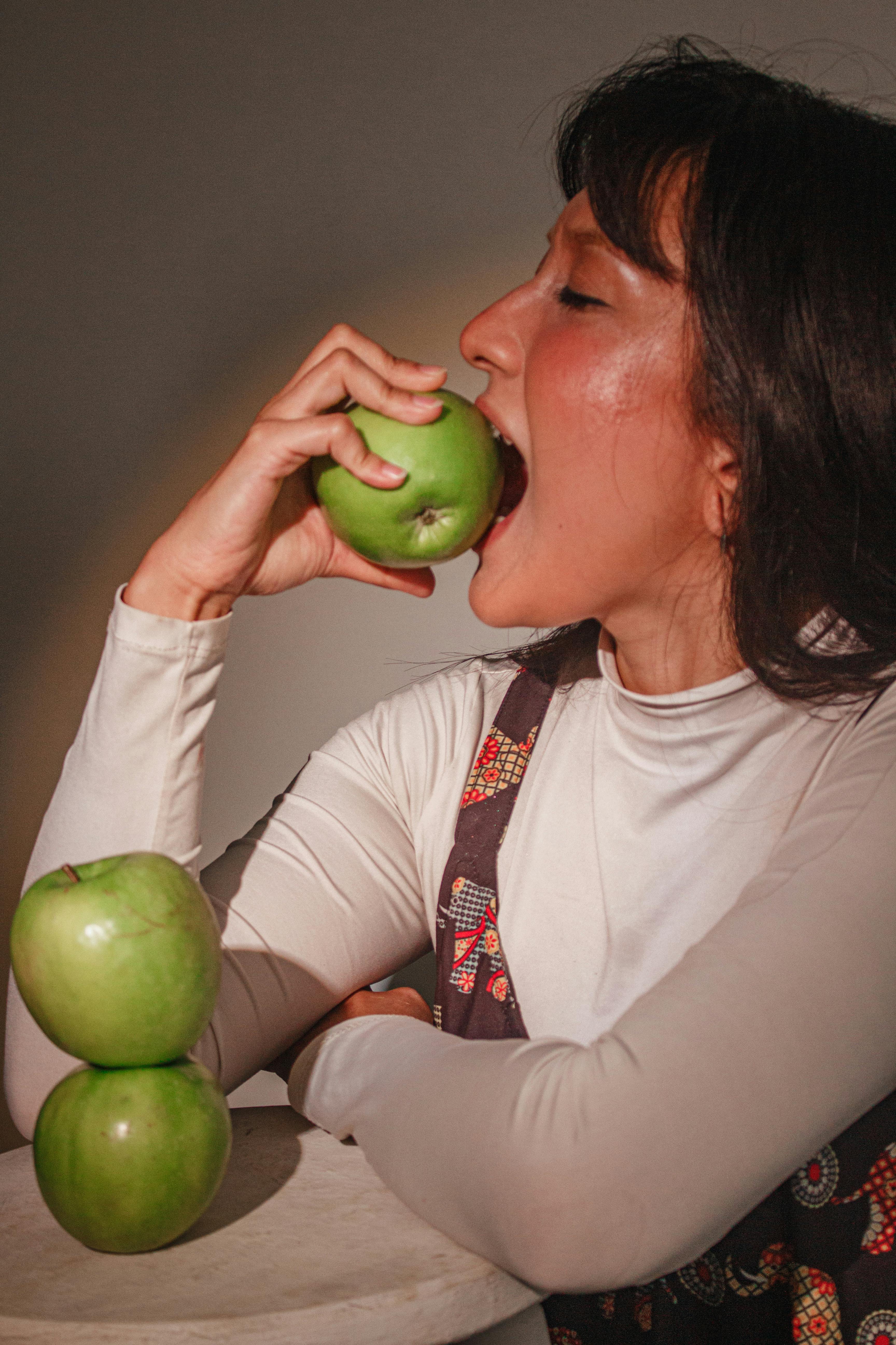 無料 鮮やかな照明の下で、積み重ねられたリンゴの中から、女性が青リンゴを一口食べています。 写真素材