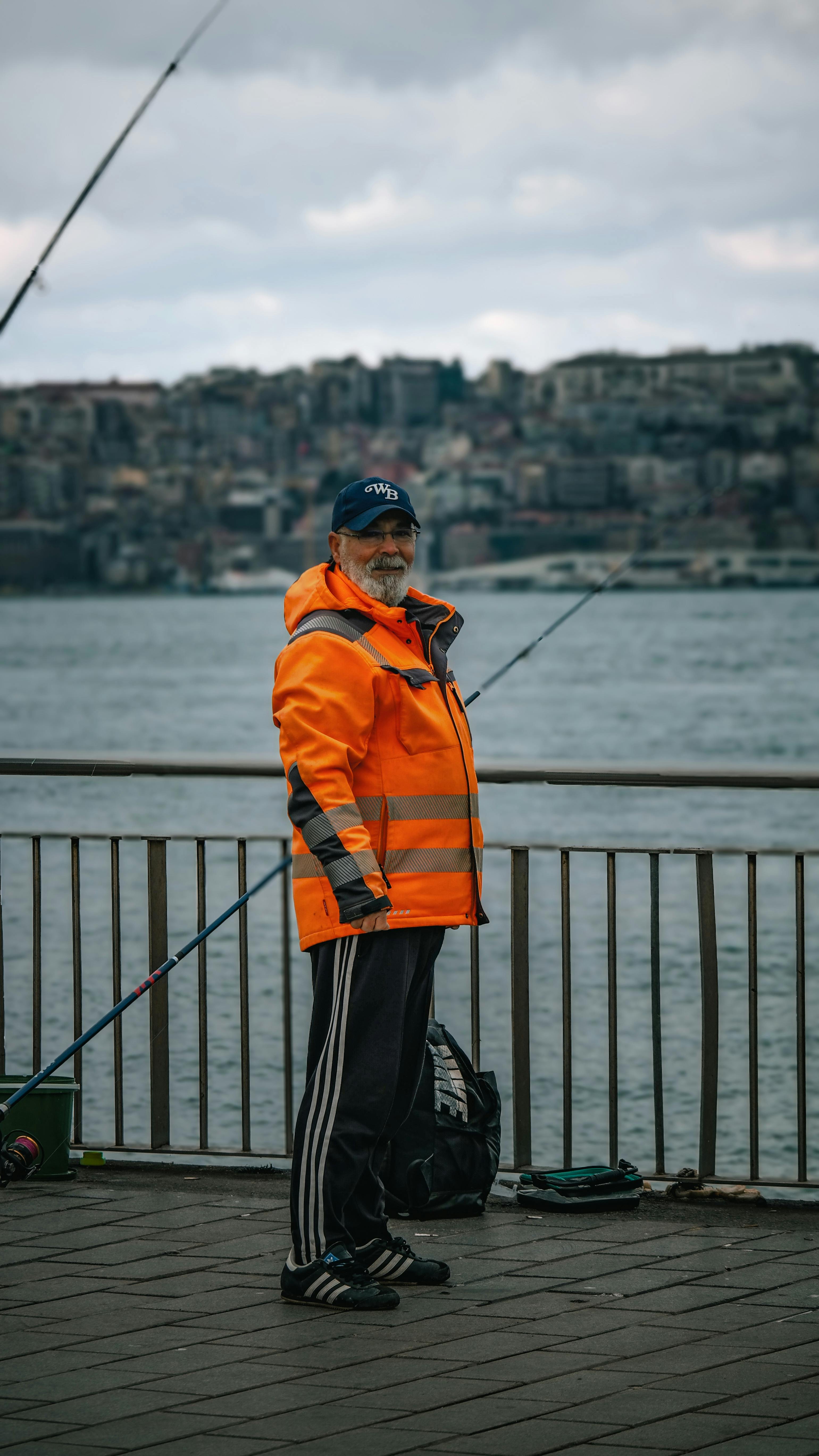 бесплатная Мужчина в оранжевой куртке ловит рыбу на городской набережной, пасмурный день. Стоковое фото