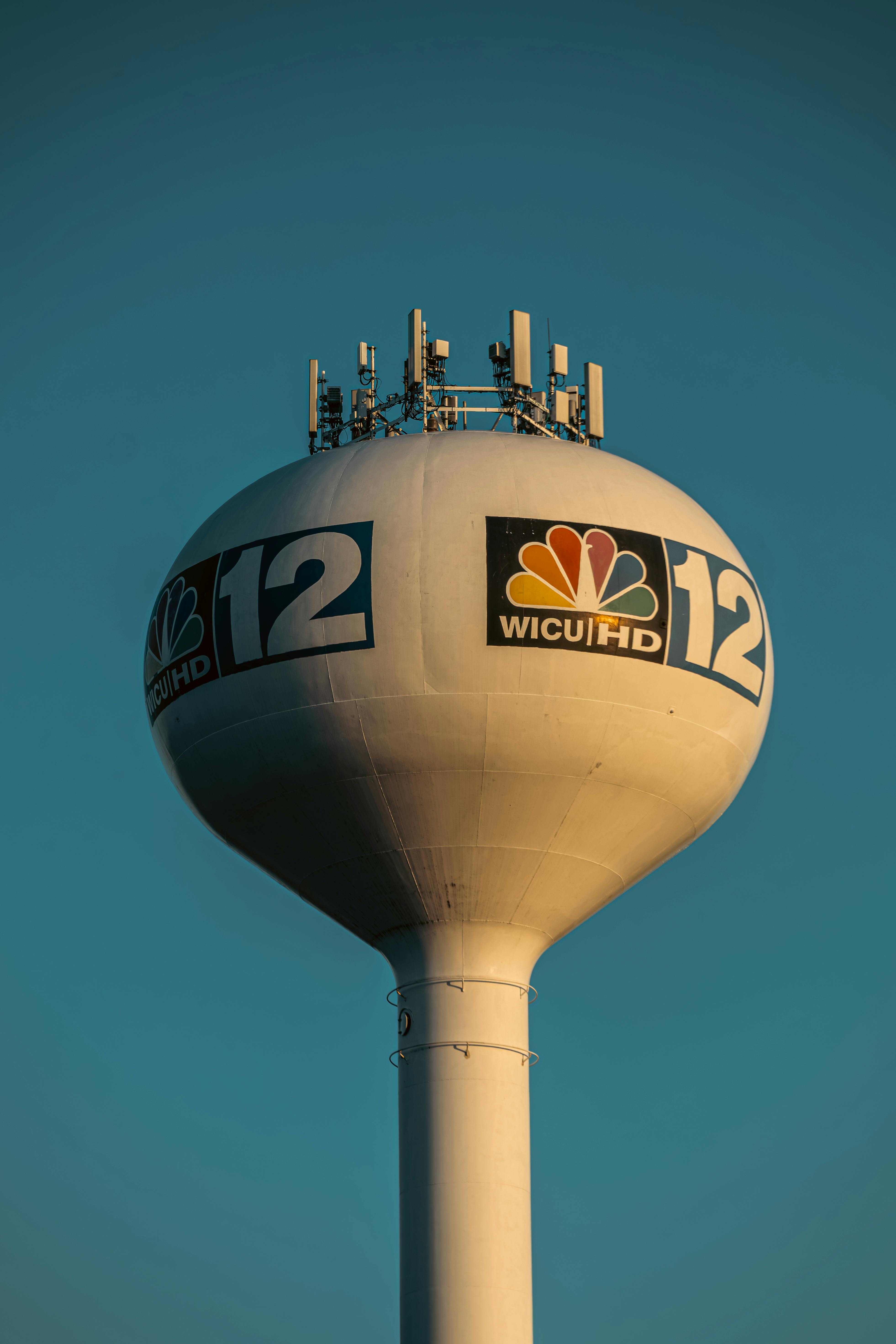 Free Water tower featuring broadcasting logos under a clear blue sky. Stock Photo