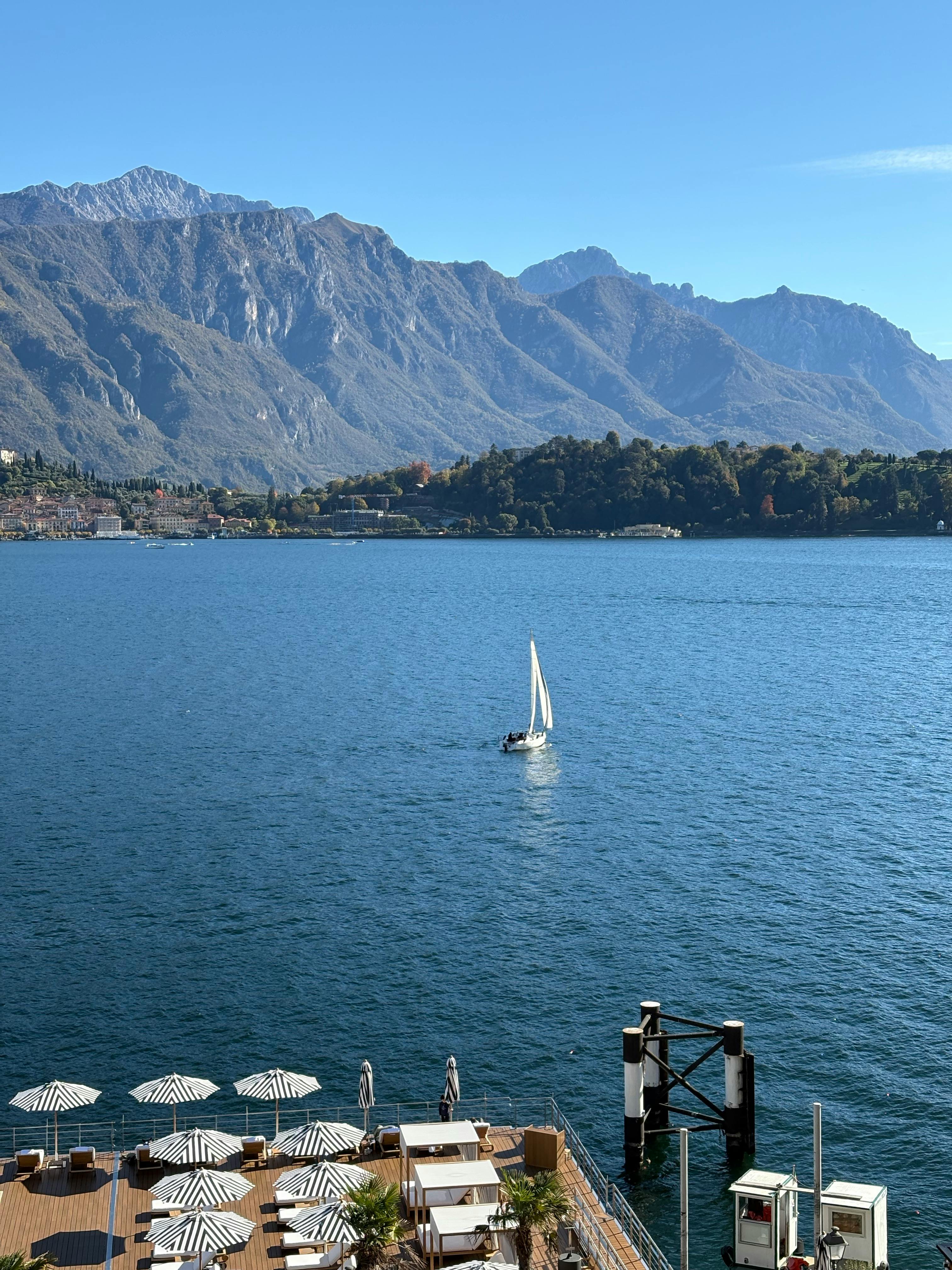 Free Tranquil lake scene with a sailboat, mountains, and a sunny terrace. Stock Photo