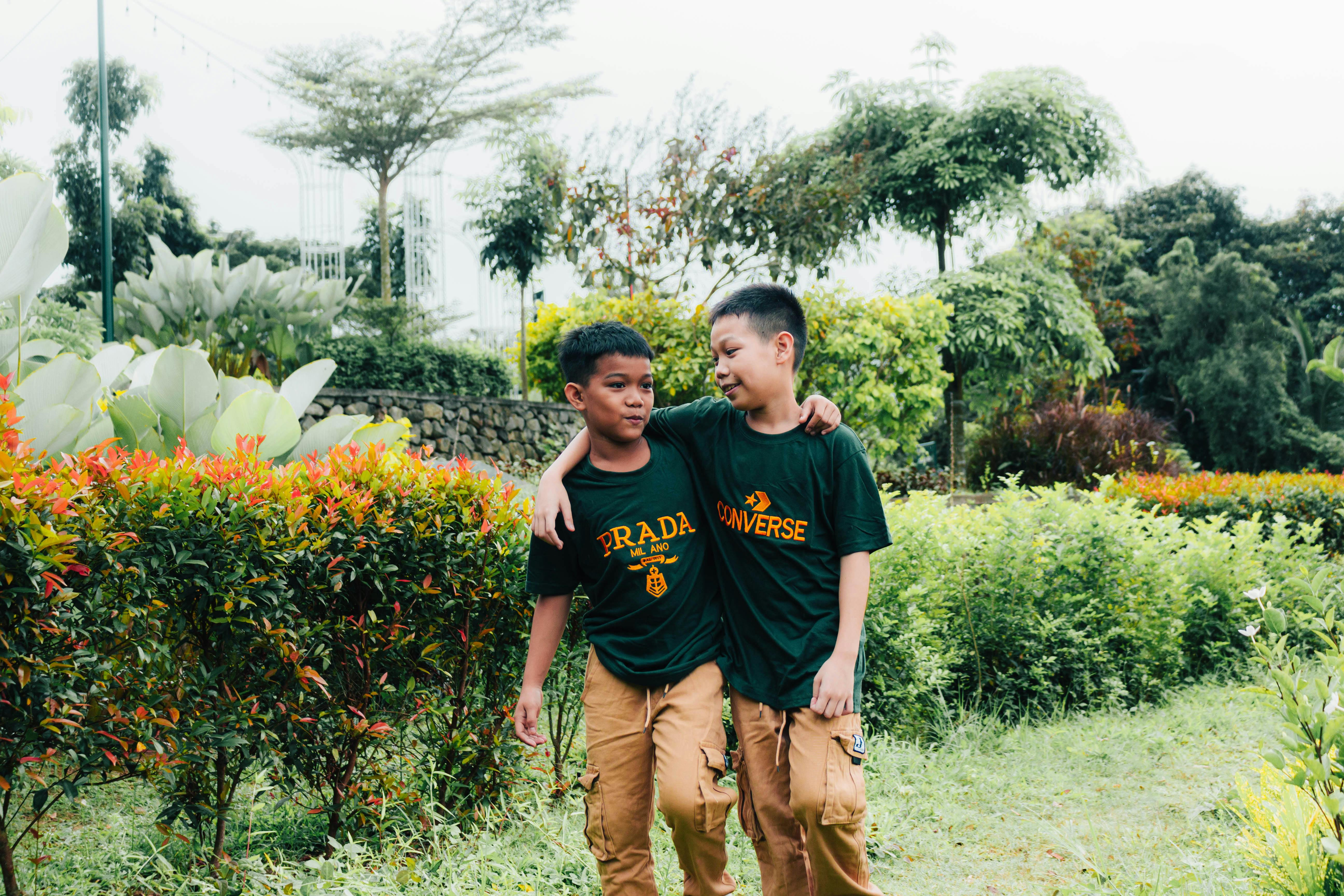 Two boys walking together in a lush garden, wearing casual outfits.