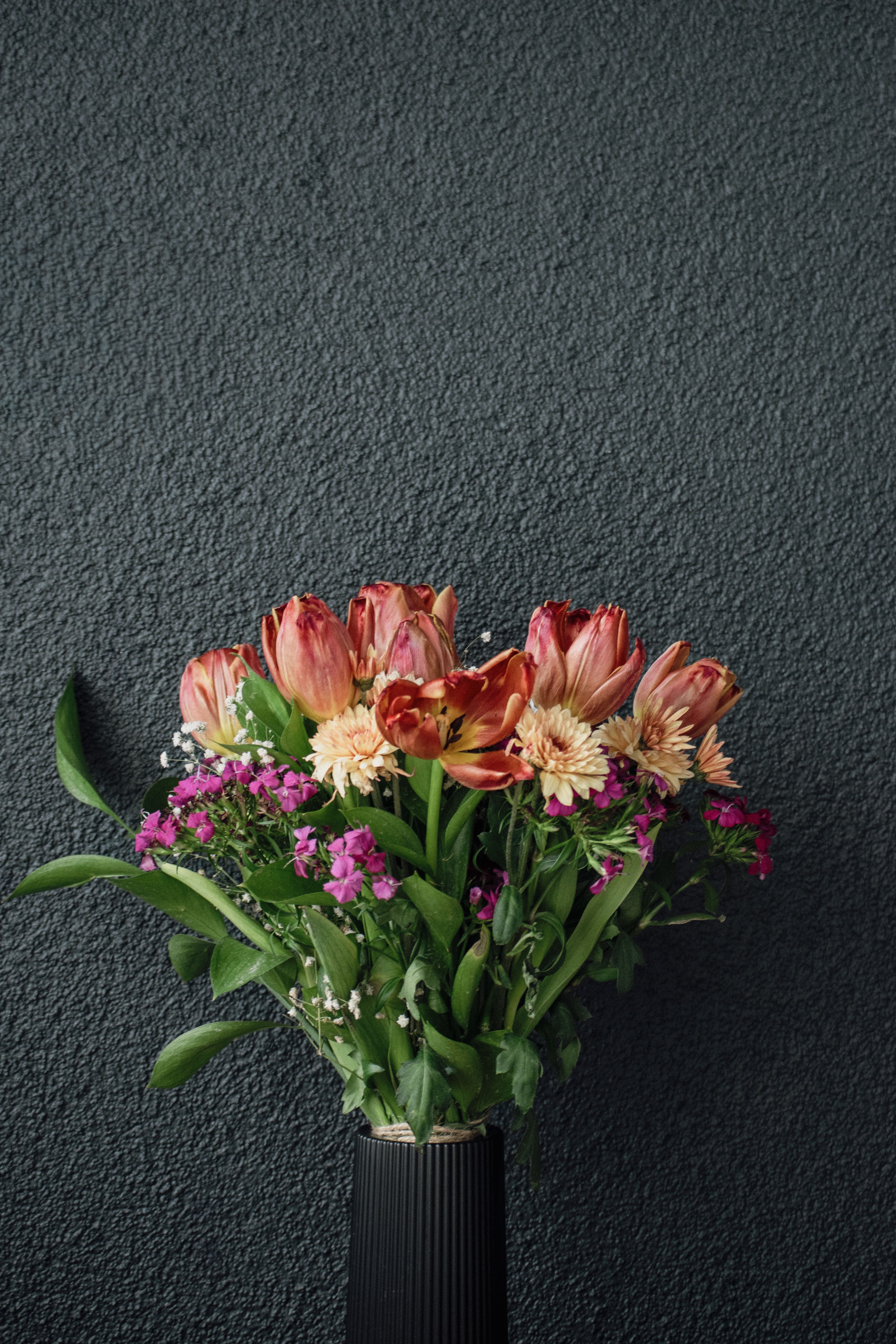 Vibrant bouquet of tulips, chrysanthemums, and wildflowers in a black vase against a textured wall.