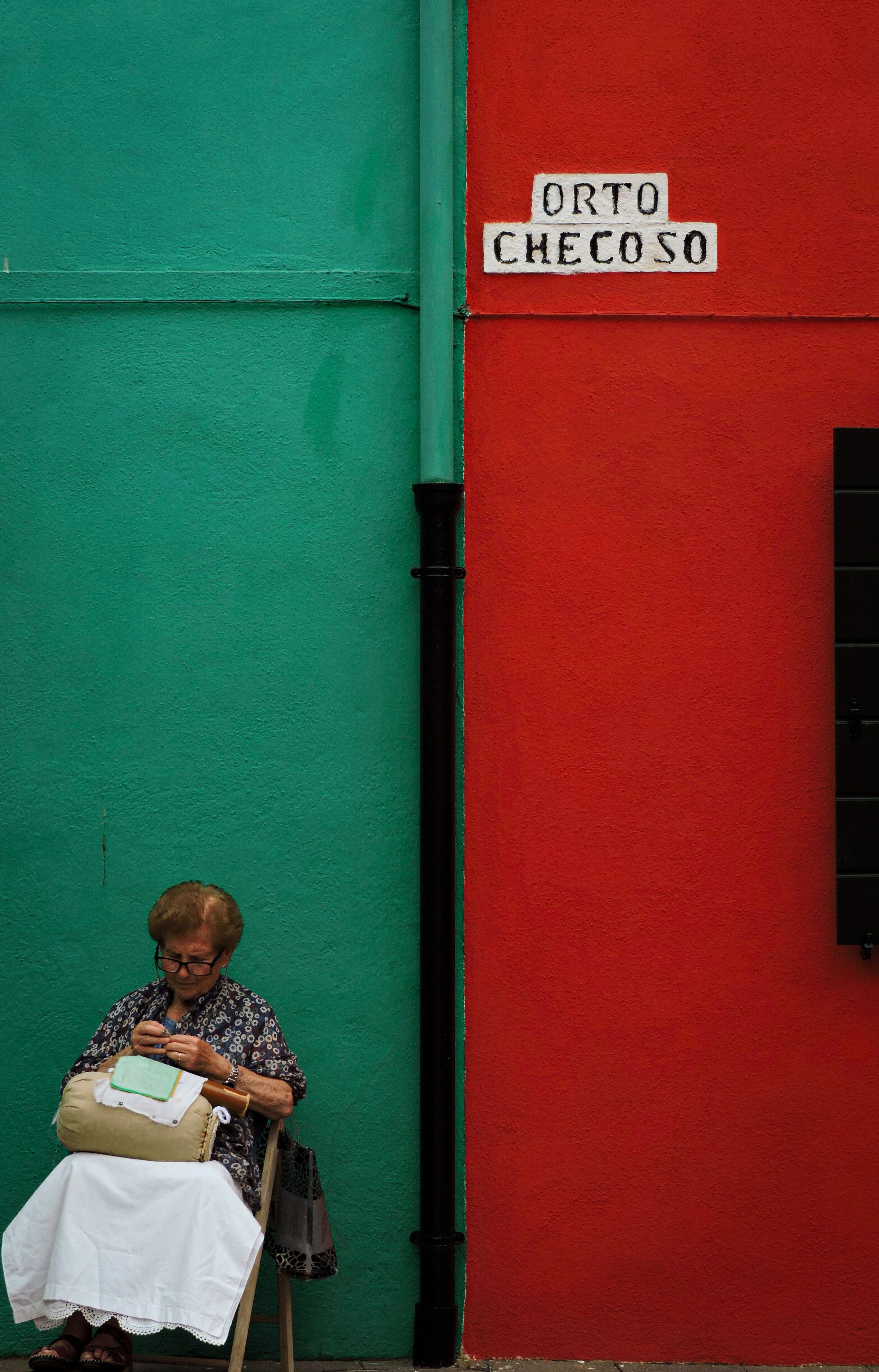 Elderly woman sewing against vibrant red and green walls in Burano, Venice.
