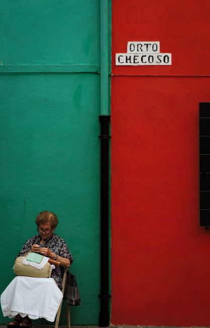 Elderly woman sewing against vibrant red and green walls in Burano, Venice.