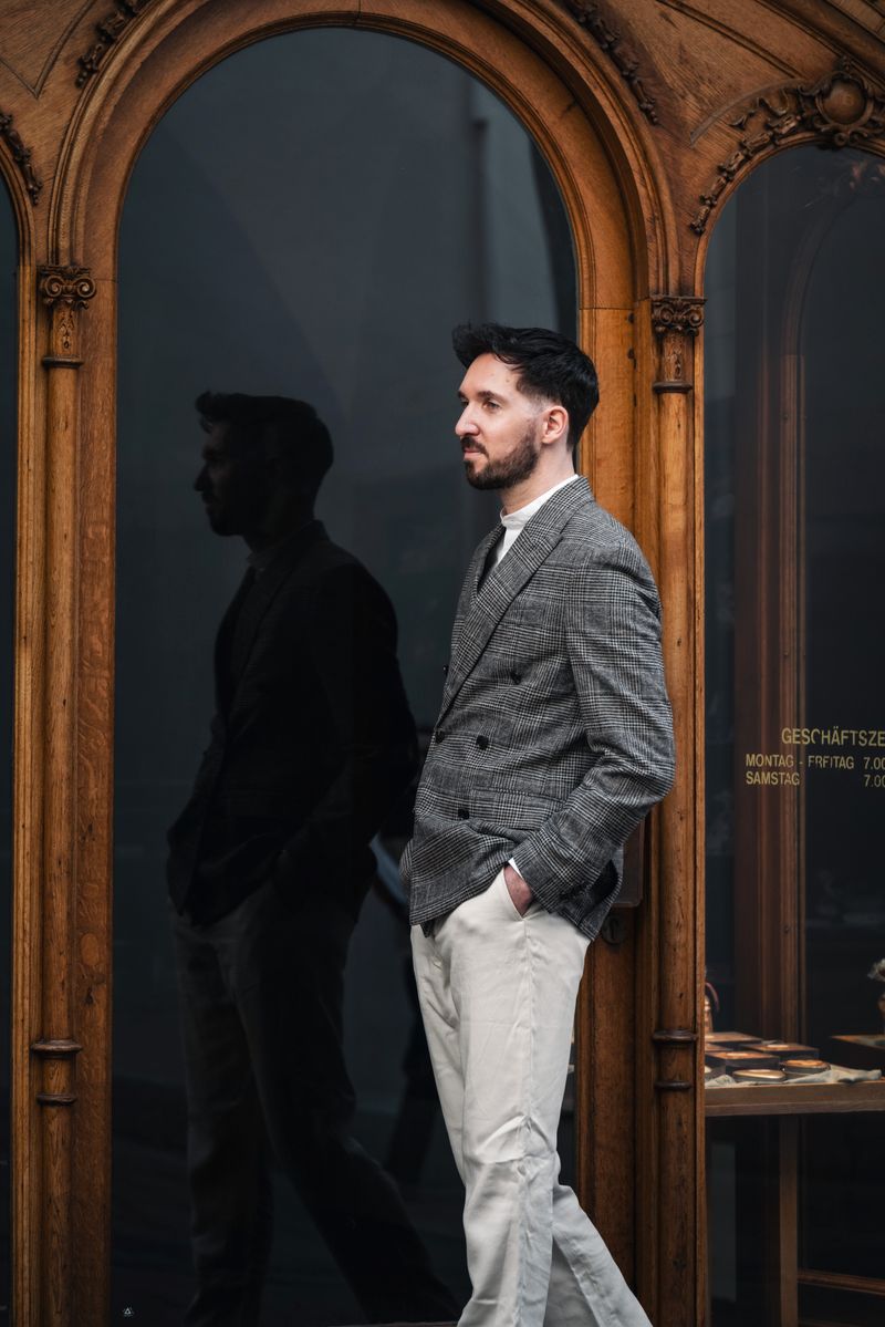 A fashionable man poses by an ornate wooden doorway in Graz, Austria, with his reflection visible.