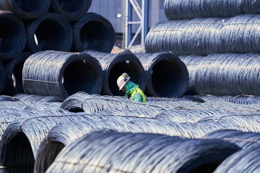 Construction worker in safety gear navigates large stacks of metal wire coils at an industrial site.