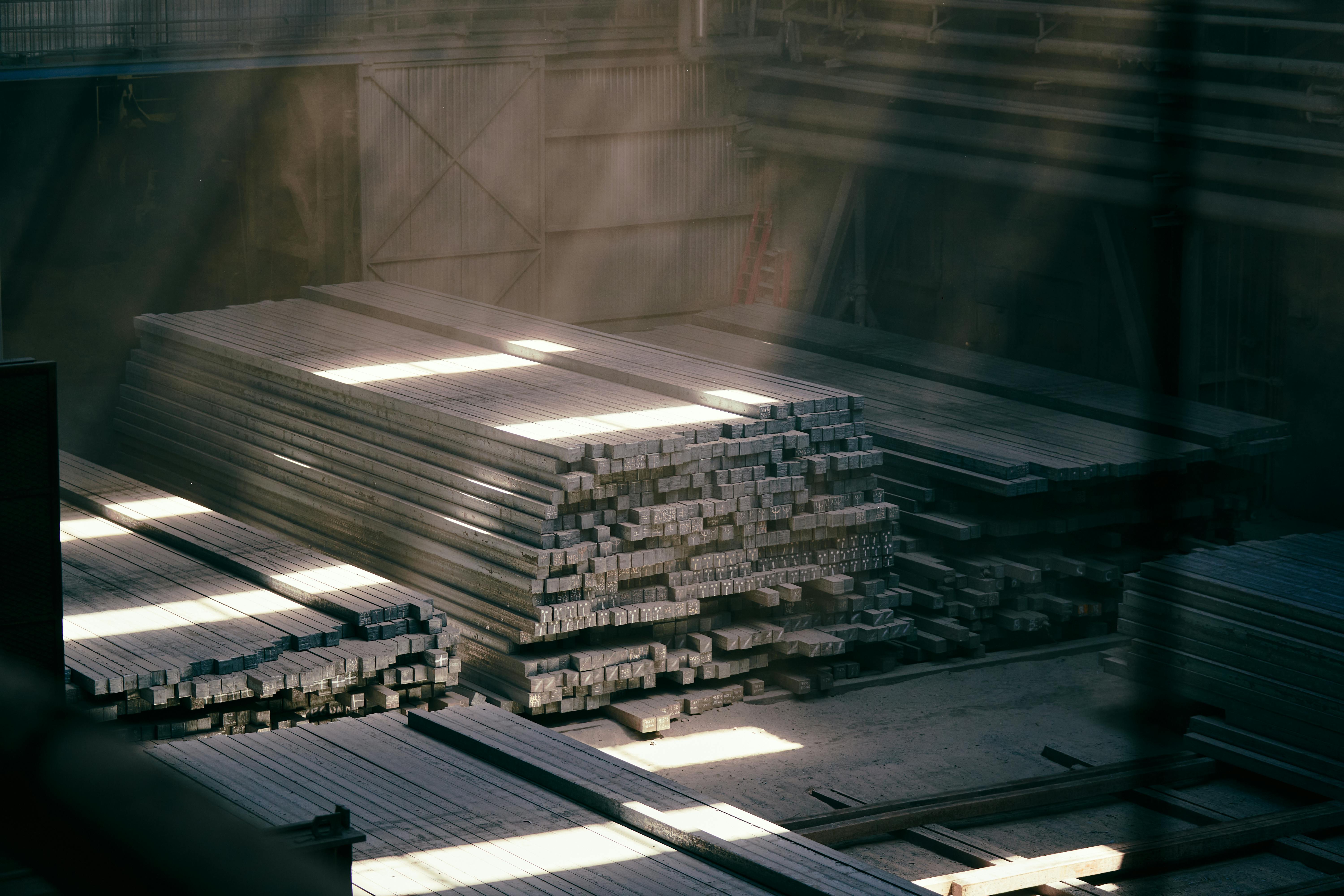 Ücretsiz Endüstriyel bir depoya süzülen güneş ışığı, üst üste yığılmış metal kirişleri aydınlatıyor. Stok Fotoğraflar