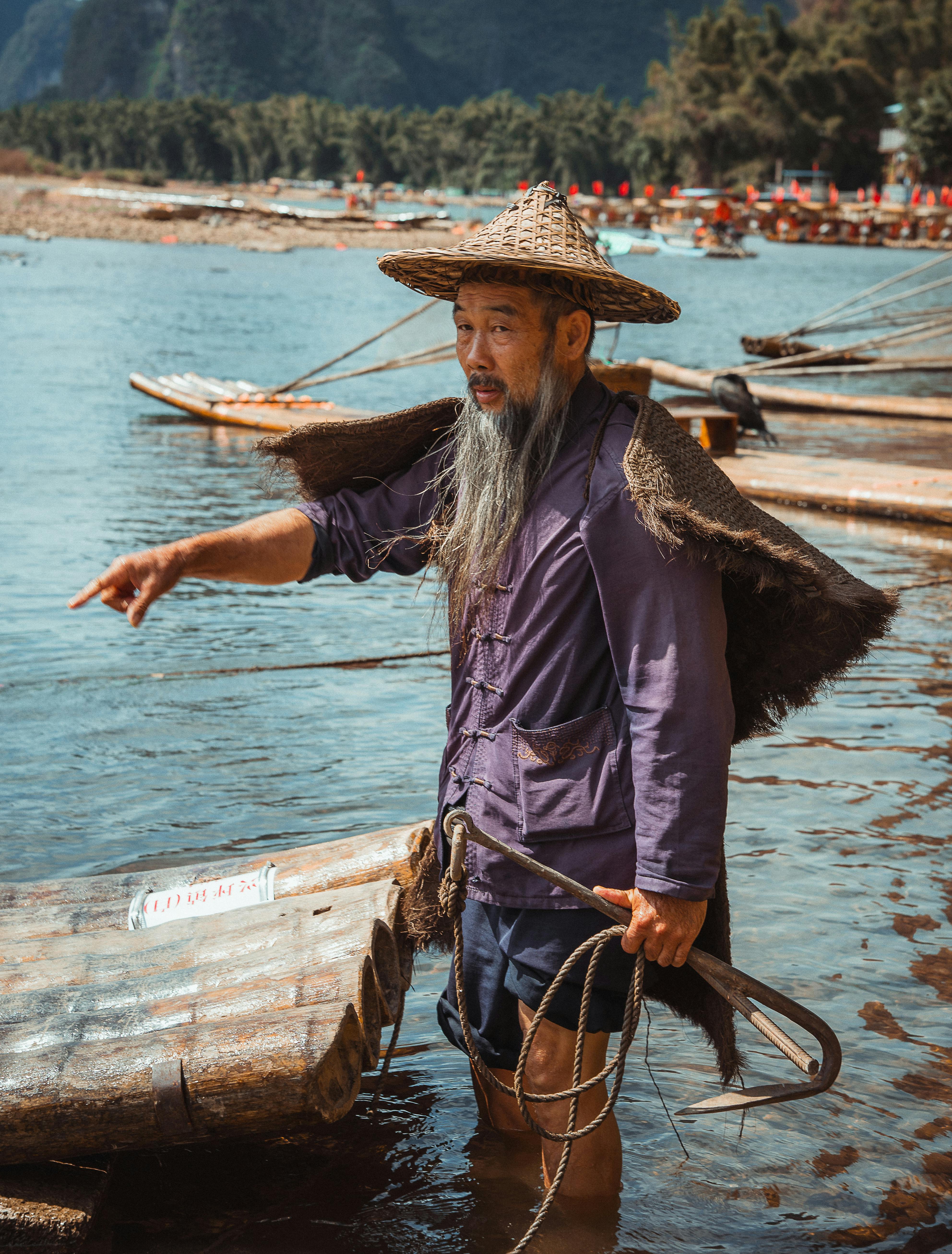 Gratis Nelayan tua bertopi jerami di Sungai Li di Guilin, Tiongkok, menunjuk ke seberang rakit kayu. Foto Stok