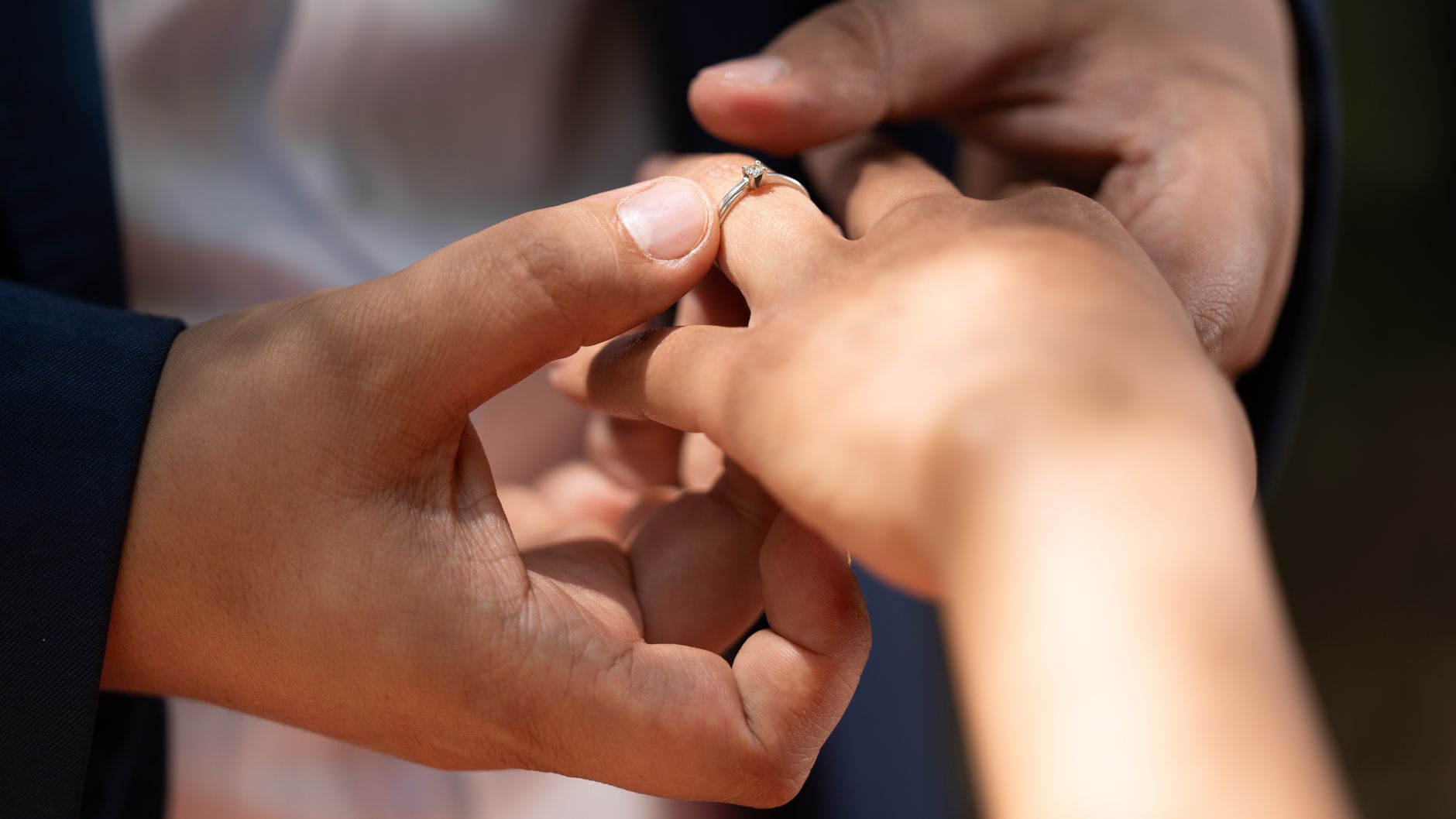 Close-up of an engagement ring being placed on a woman's hand in natural lighting.