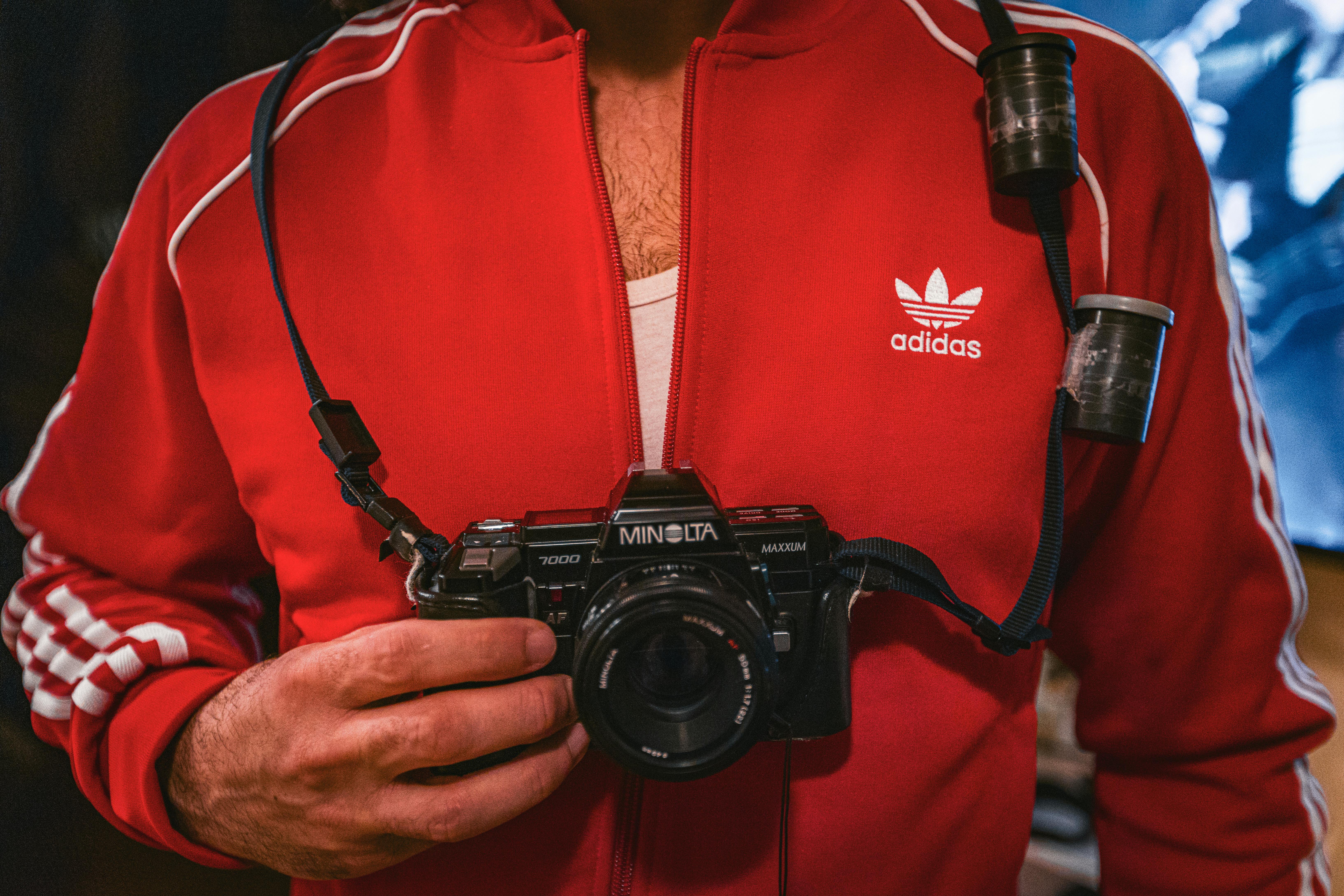 Gratis Uomo che tiene in mano una macchina fotografica Minolta d'epoca e indossa una giacca rossa Adidas in un ambiente interno. Foto a disposizione