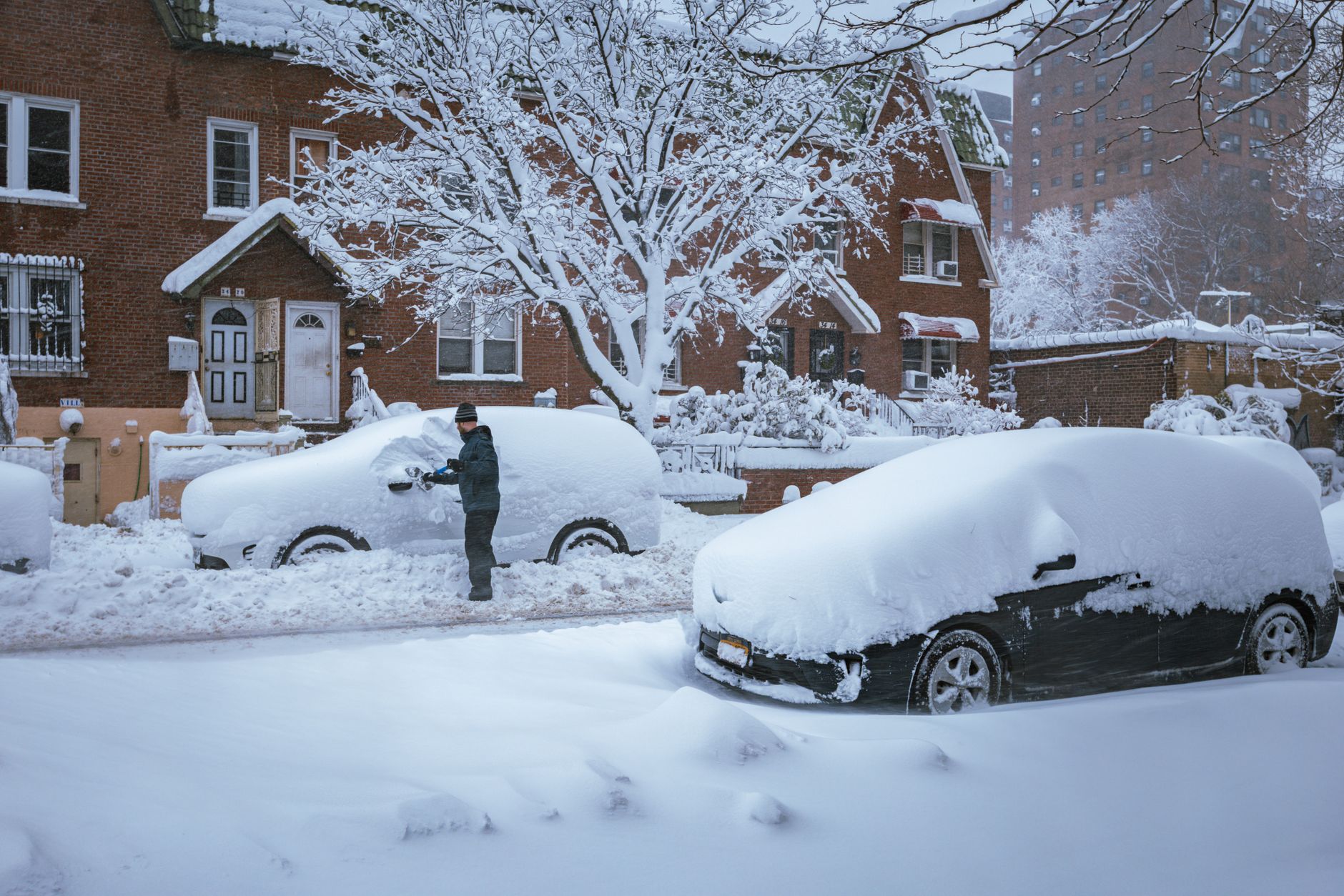 A person clears snow off a car in a snow-covered residential neighborhood after a storm.