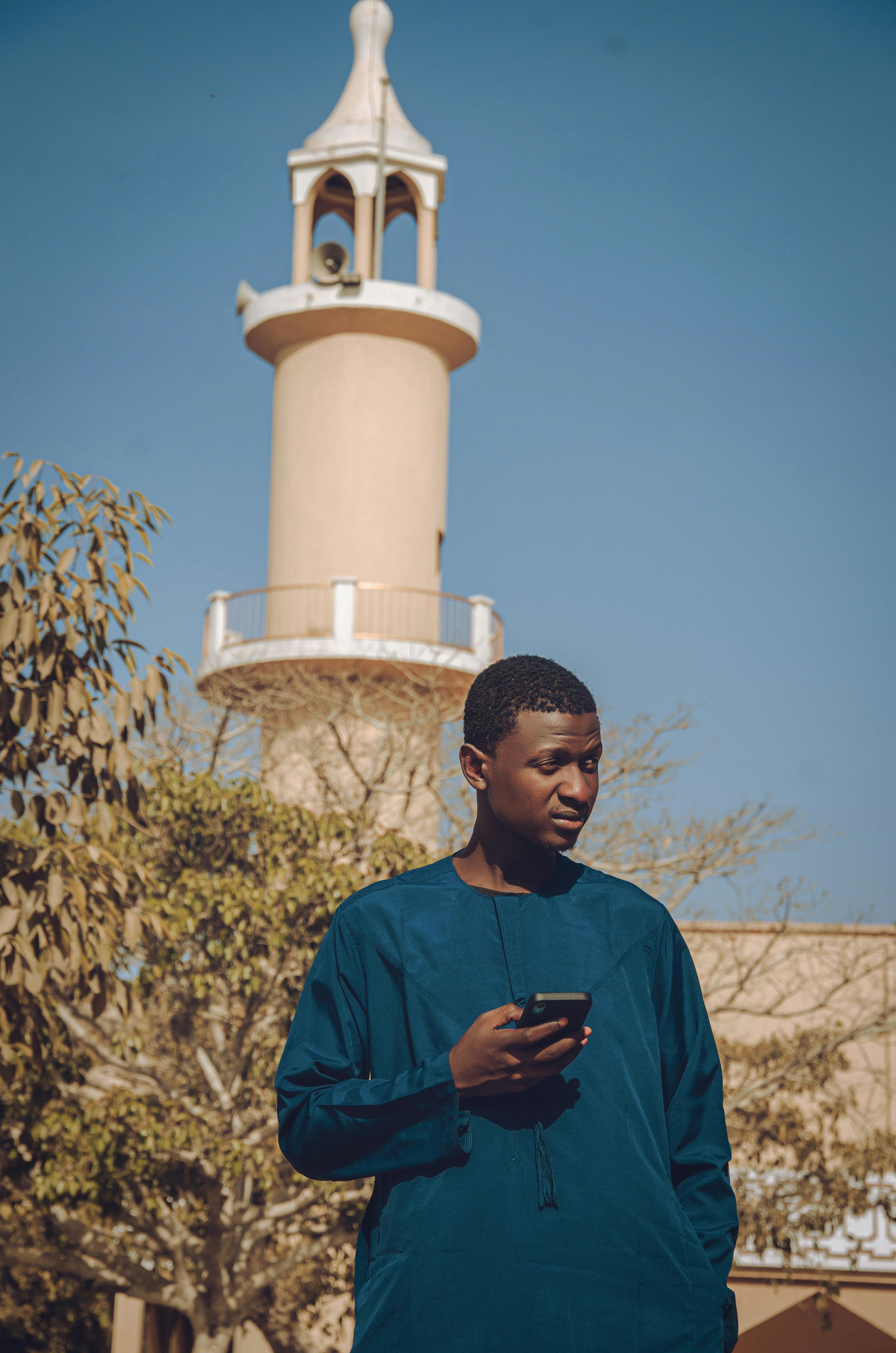 Free Young man holding smartphone standing near mosque tower on a sunny day. Stock Photo
