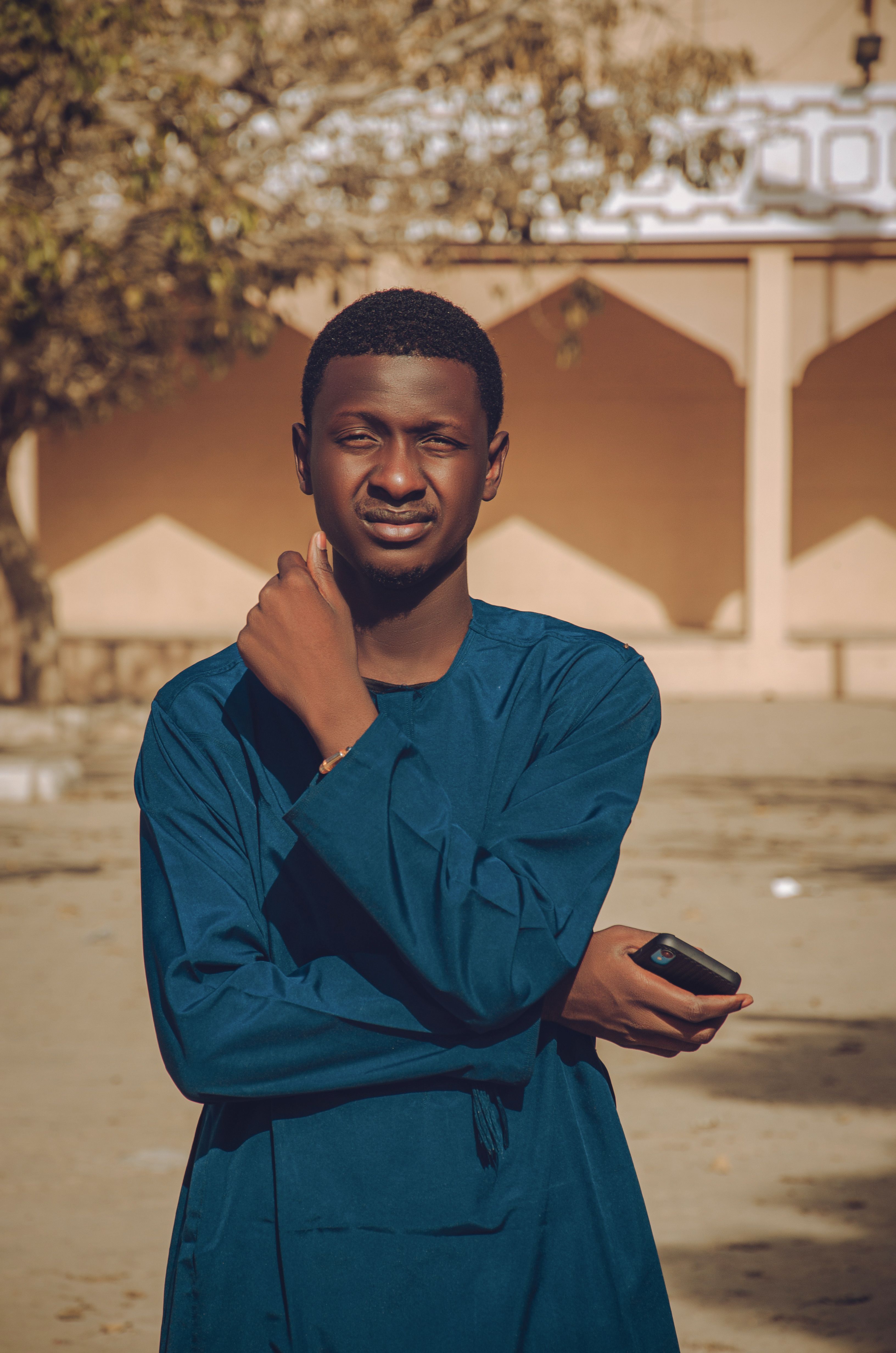 Young man in blue traditional attire stands outdoors holding a phone.