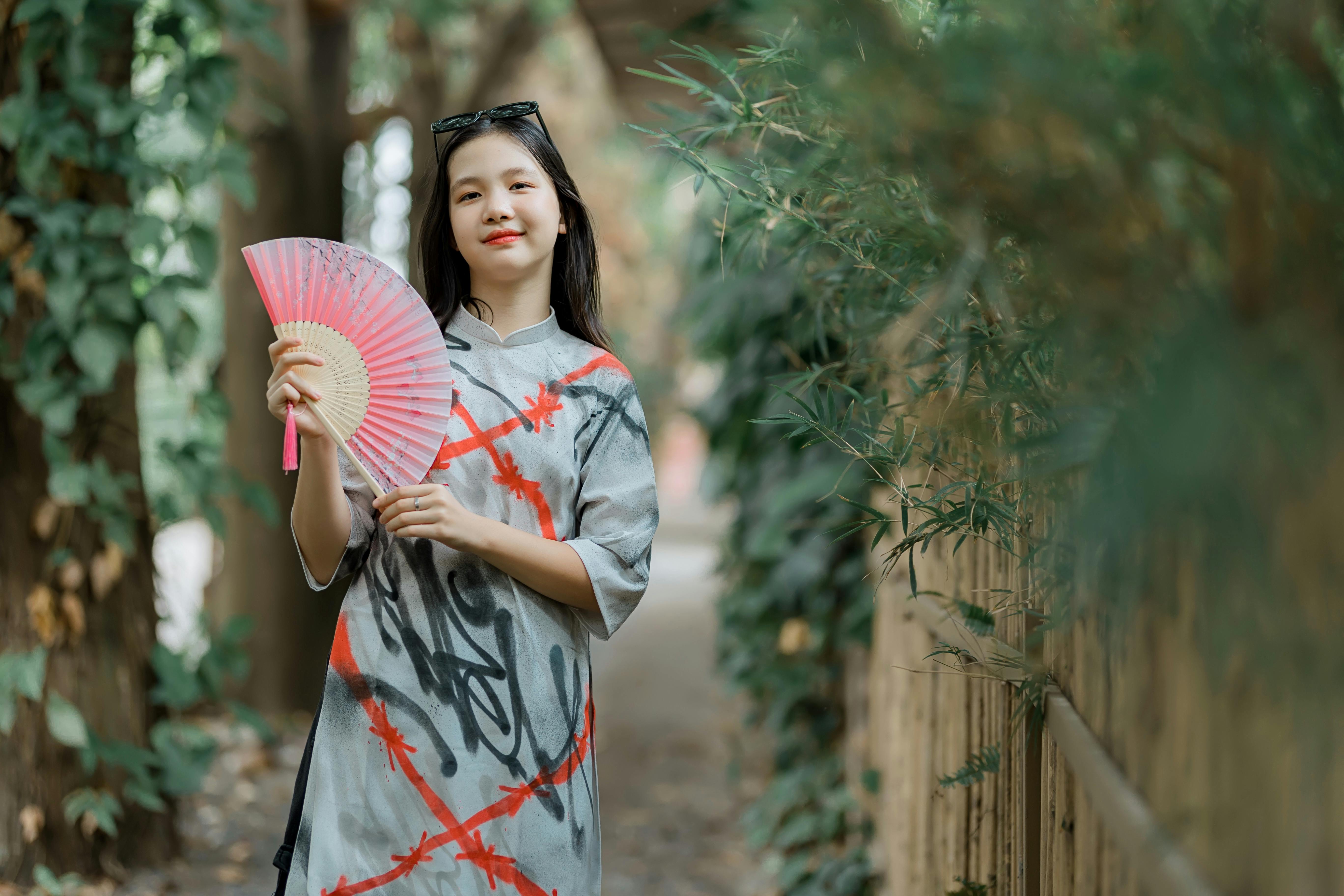 Free Young woman in a colorful garment holding a bamboo hand fan, enjoying a peaceful outdoor setting. Stock Photo