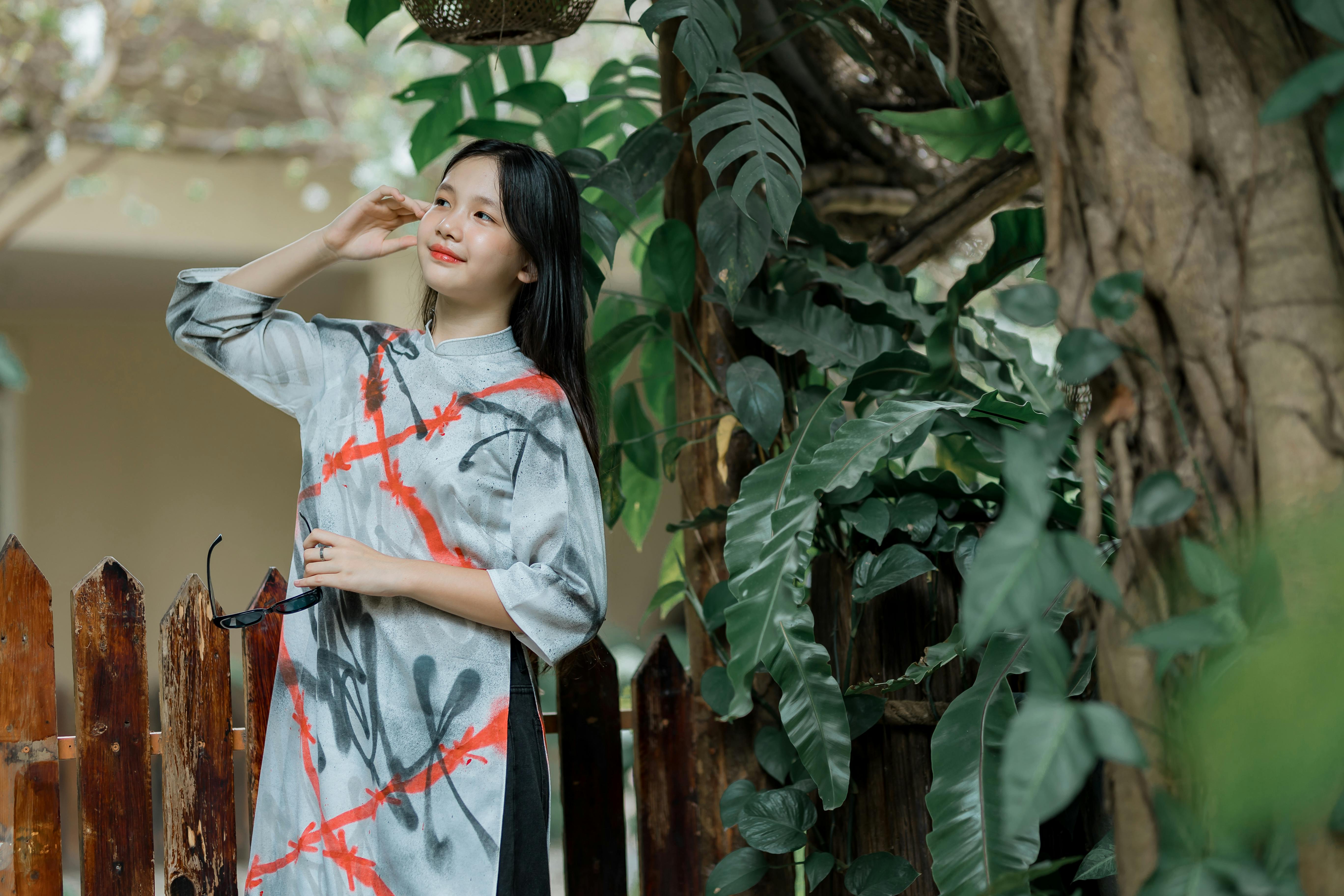 бесплатная Очаровательная молодая женщина в современном платье ао дай стоит среди пышной зелени и деревянного забора. Стоковое фото