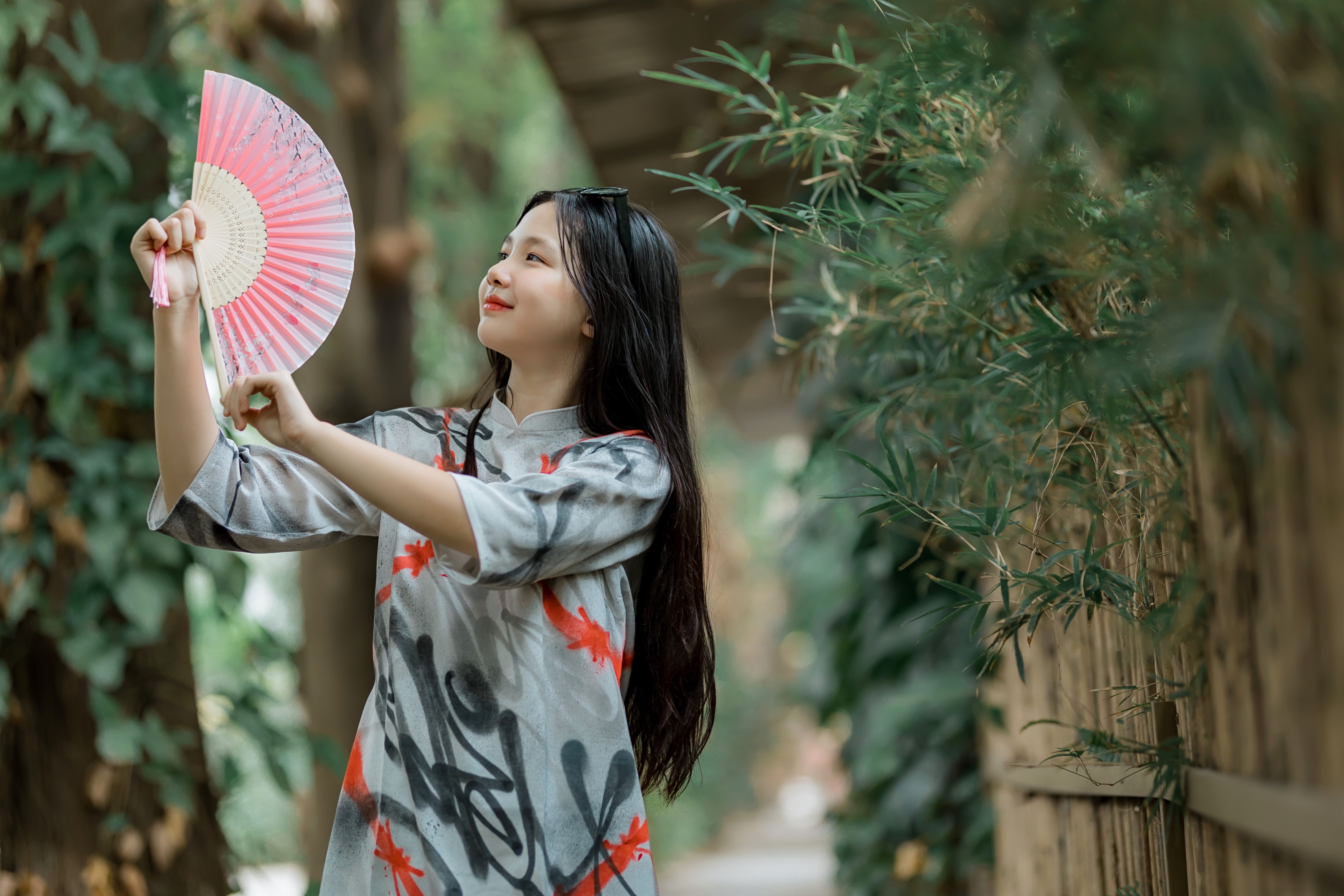 A young woman in traditional attire holding a pink fan in a serene outdoor setting.