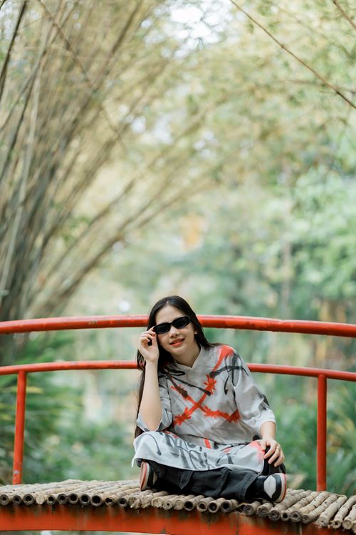 Free Woman in sunglasses relaxing outdoors on a red bridge surrounded by lush greenery. Stock Photo