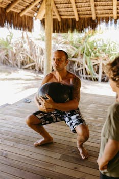A man exercises with a medicine ball on a wooden deck under a thatched roof in daylight.