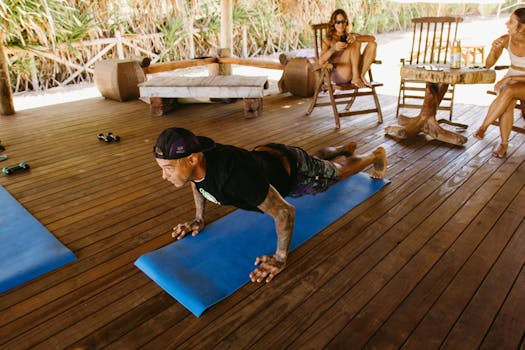 A man performs planks on a yoga mat outdoors while others relax, enjoying the natural setting.