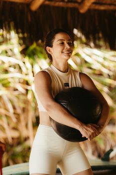 Cheerful woman holding a medicine ball during a fitness training session in a tropical setting.