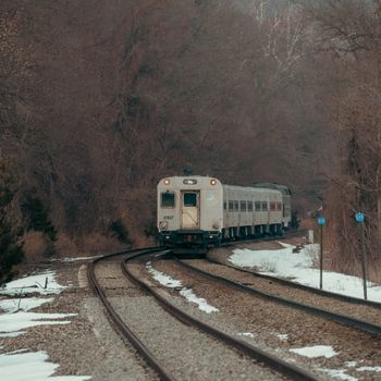 A commuter train navigates through a quiet, snow-dusted forest with bare trees, captured during winter.