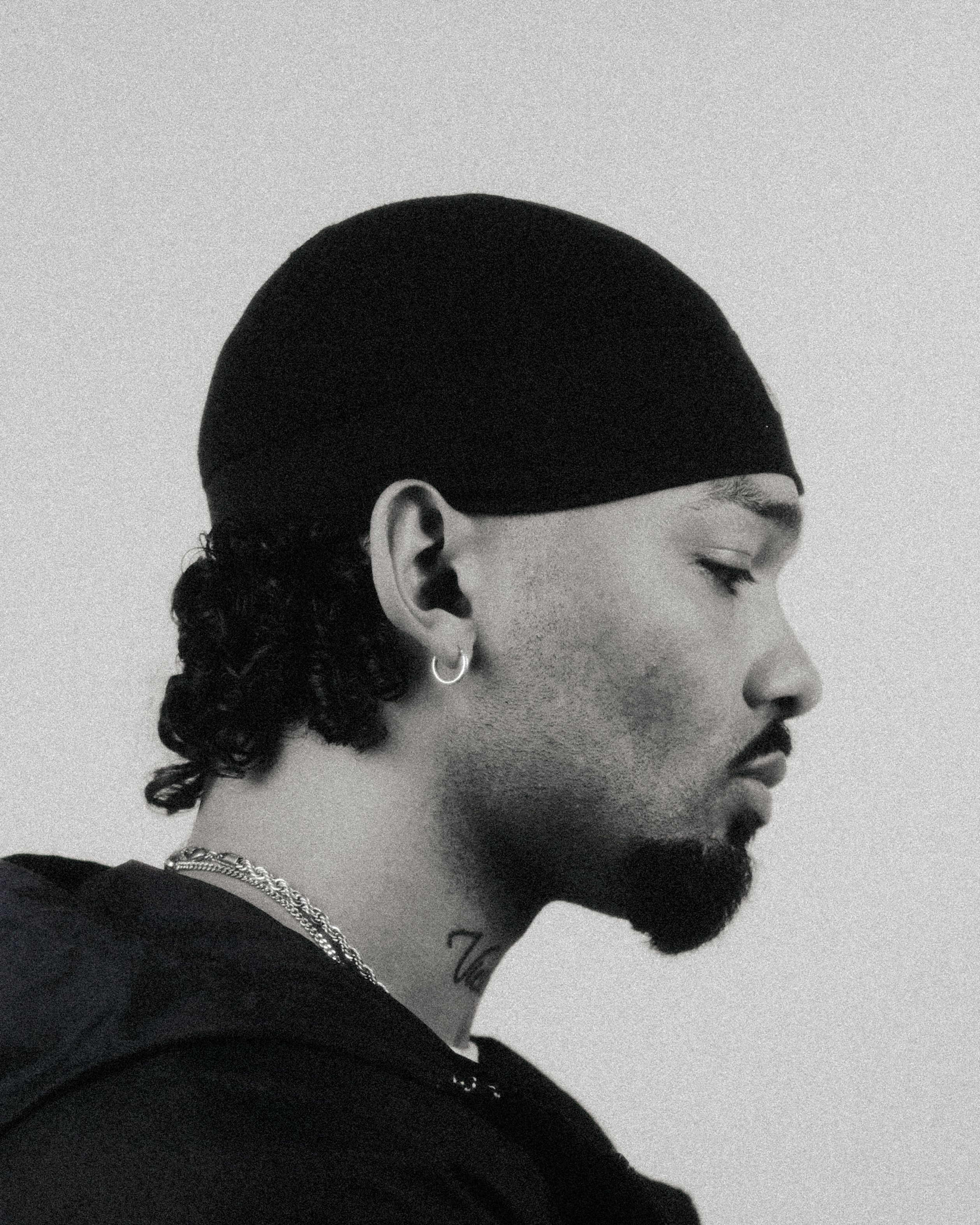 Artistic black and white profile portrait of a man wearing a beanie and jewelry.