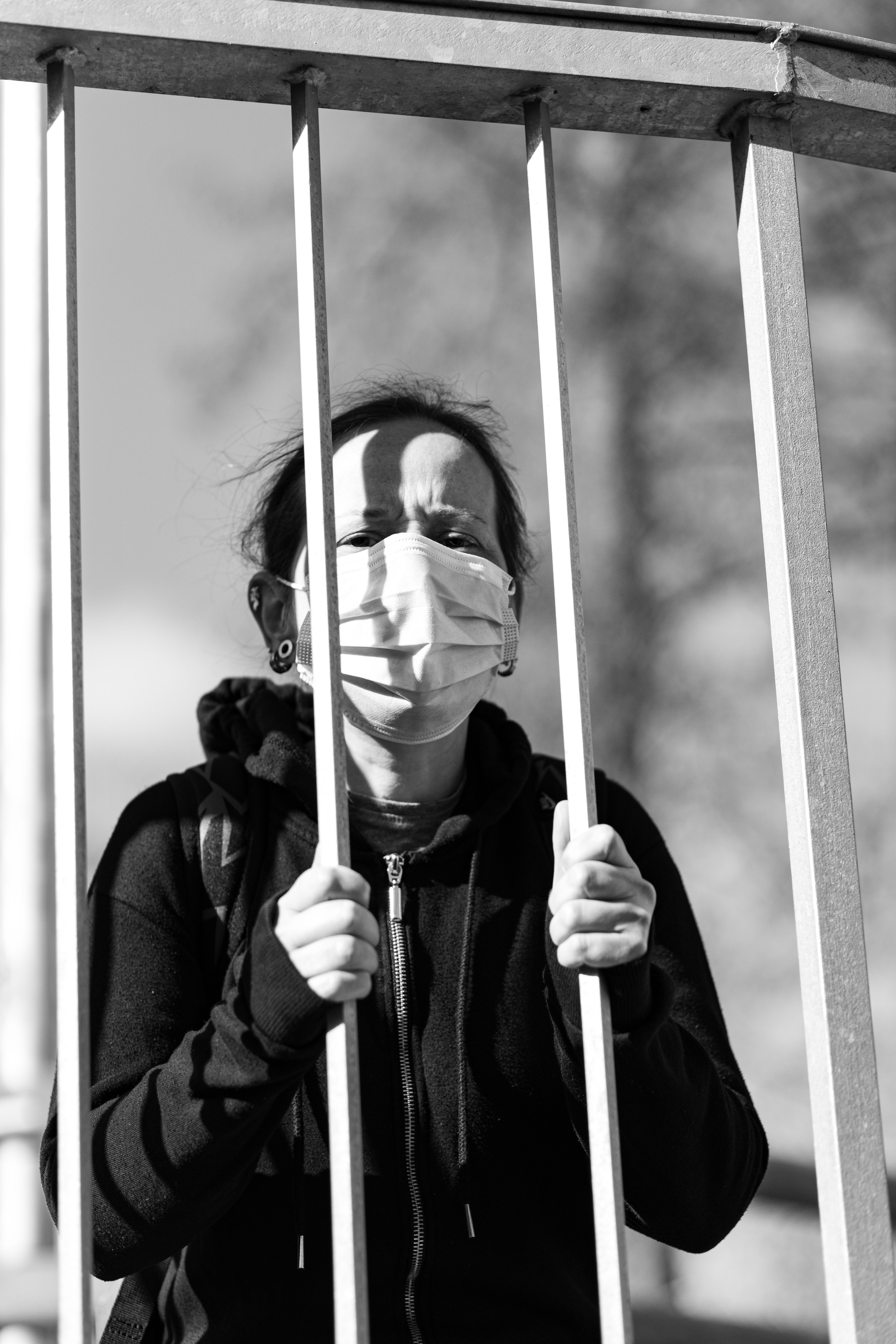 Black and white portrait of a woman wearing a face mask standing behind a metal fence outdoors.