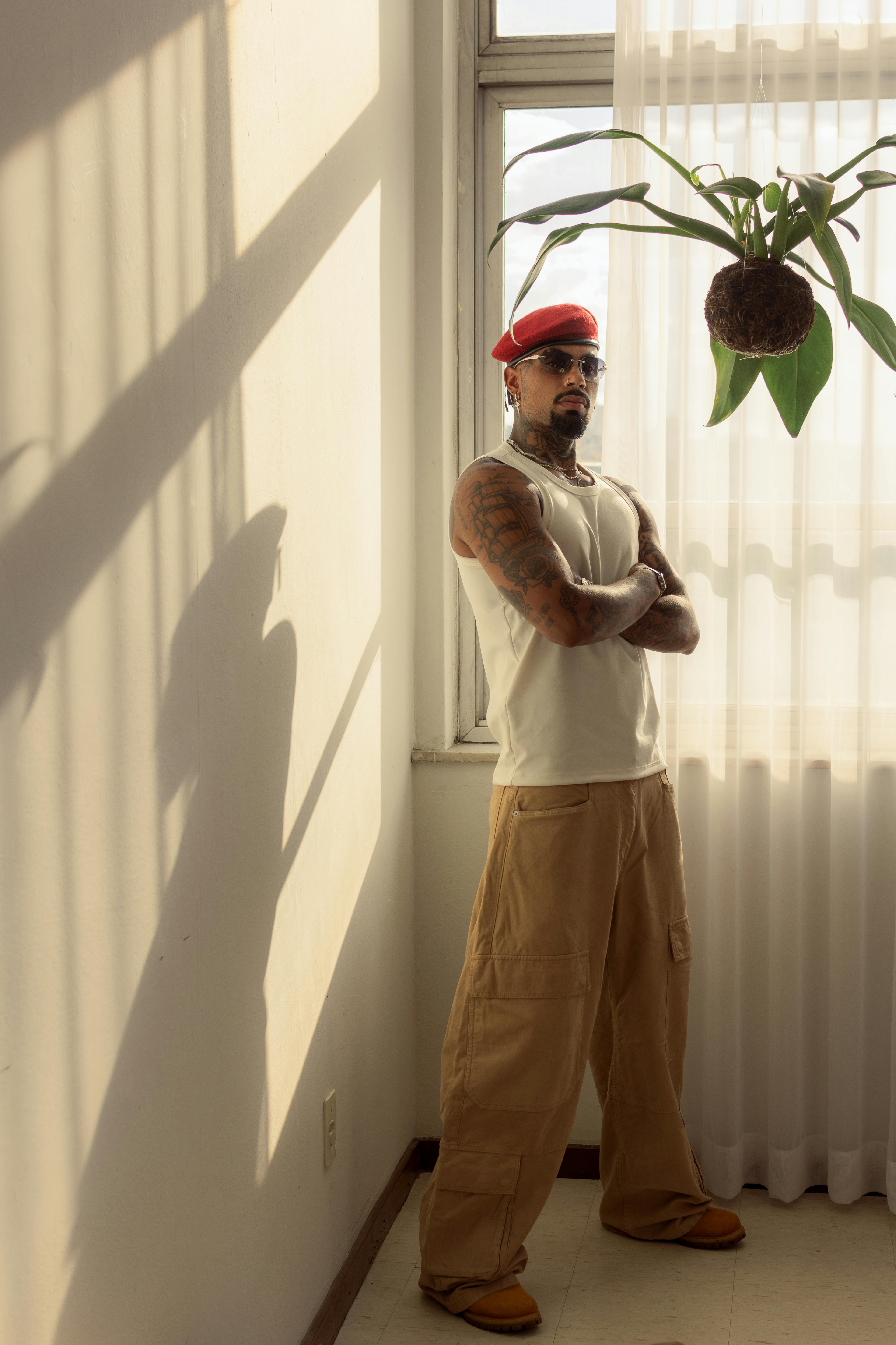 бесплатная Модный мужчина в красном берете и повседневной одежде стоит у окна, скрестив руки. Стоковое фото