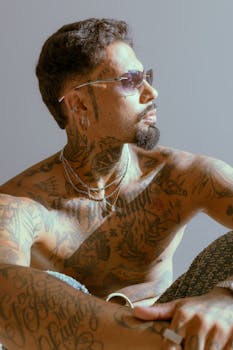 A stylish man with tattoos and sunglasses in a thoughtful pose.