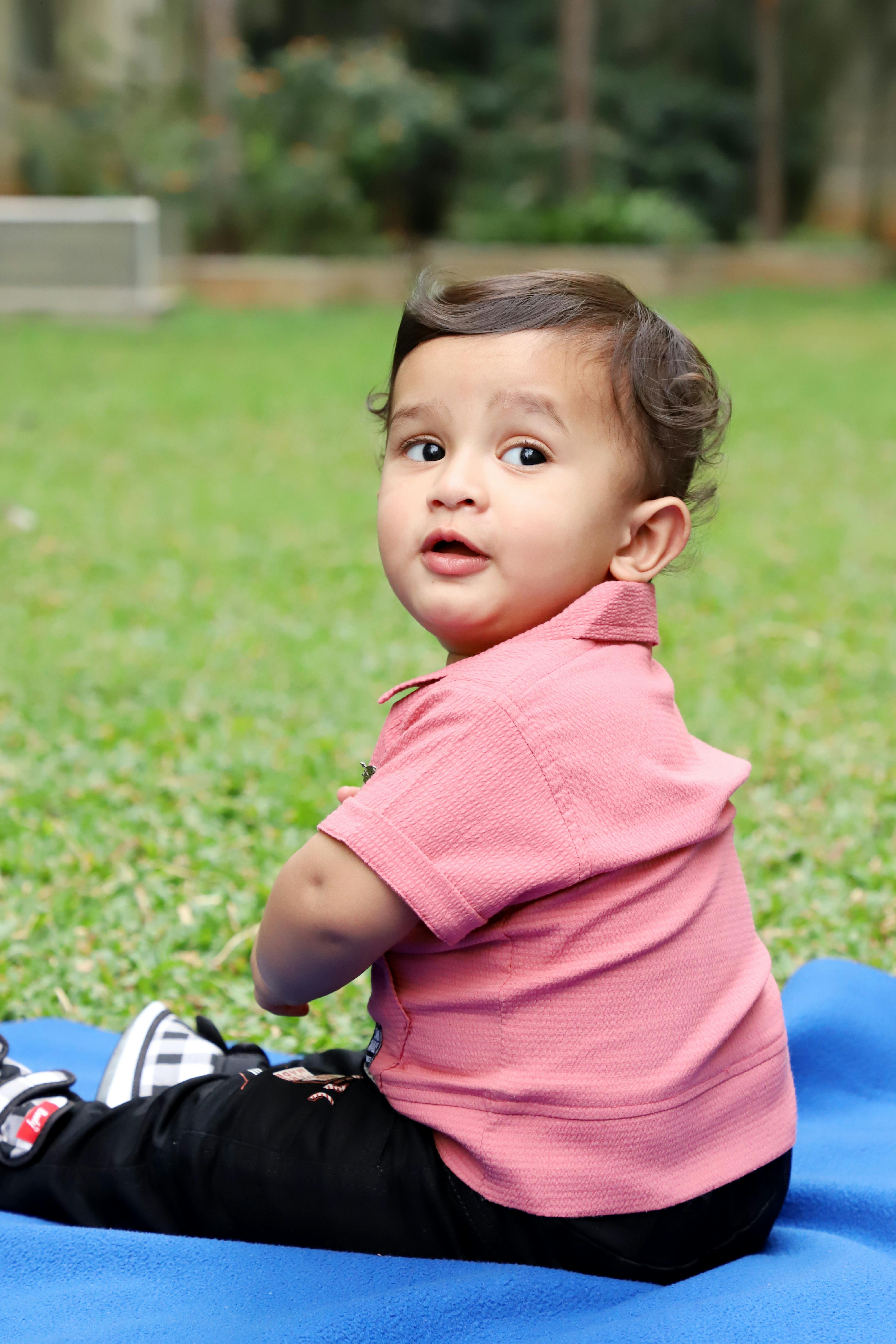 Gratis Balita menggemaskan berbaju merah muda duduk di atas selimut biru di rumput hijau, menoleh ke belakang. Foto Stok