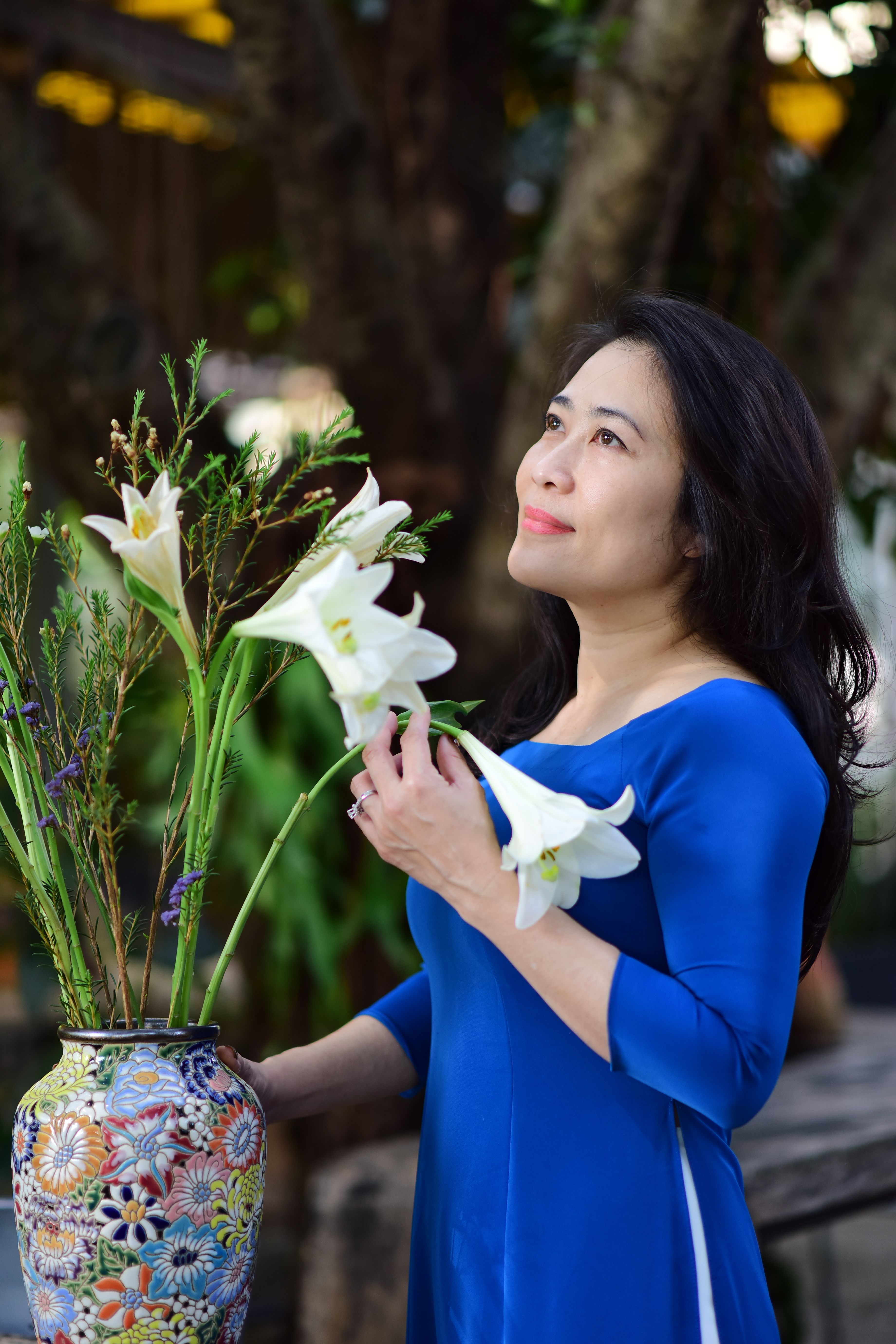 Elegant woman in blue dress holding lilies in a decorative vase outdoors.