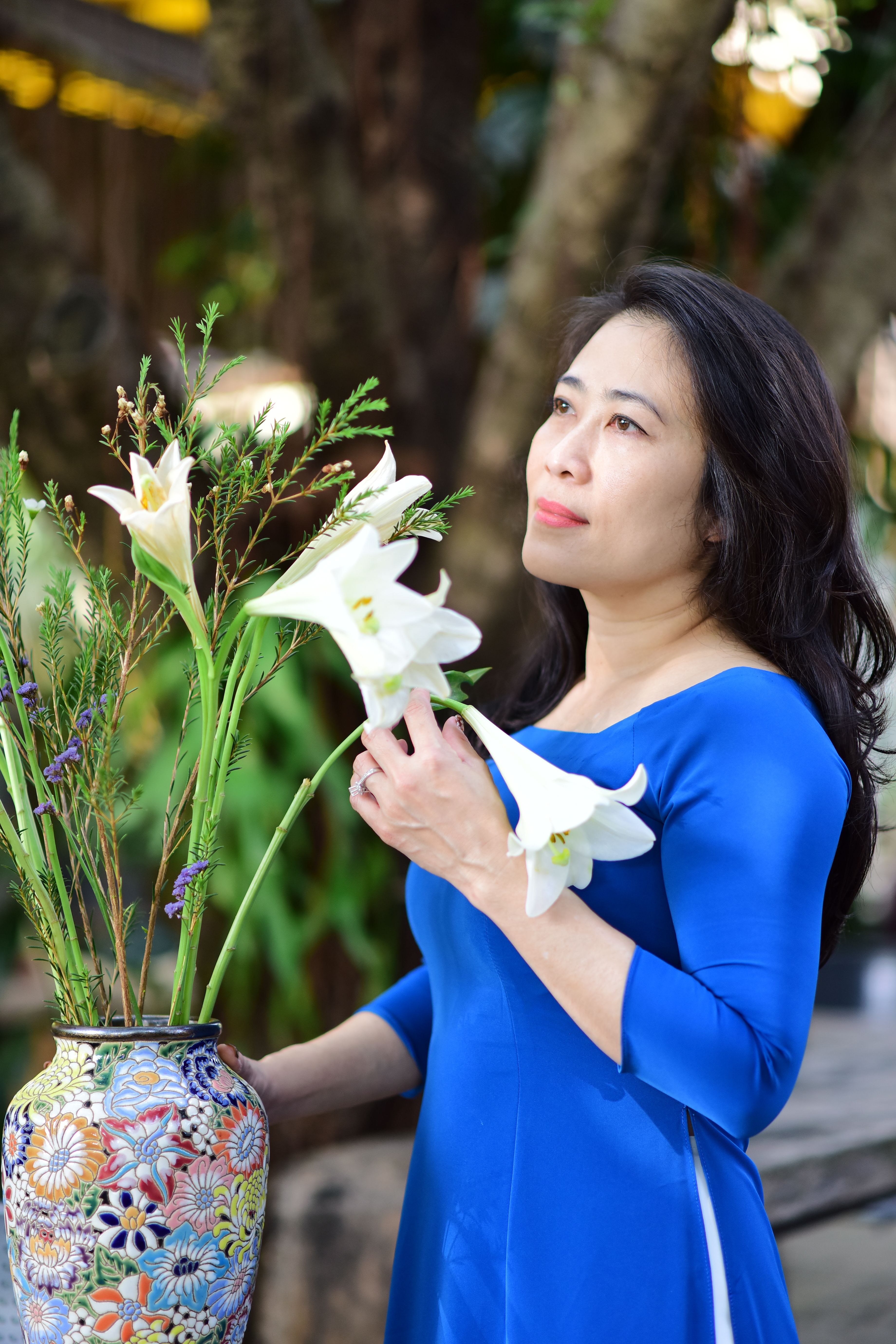 Elegant woman in blue arranging white lilies in a colorful vase in a serene outdoor setting.