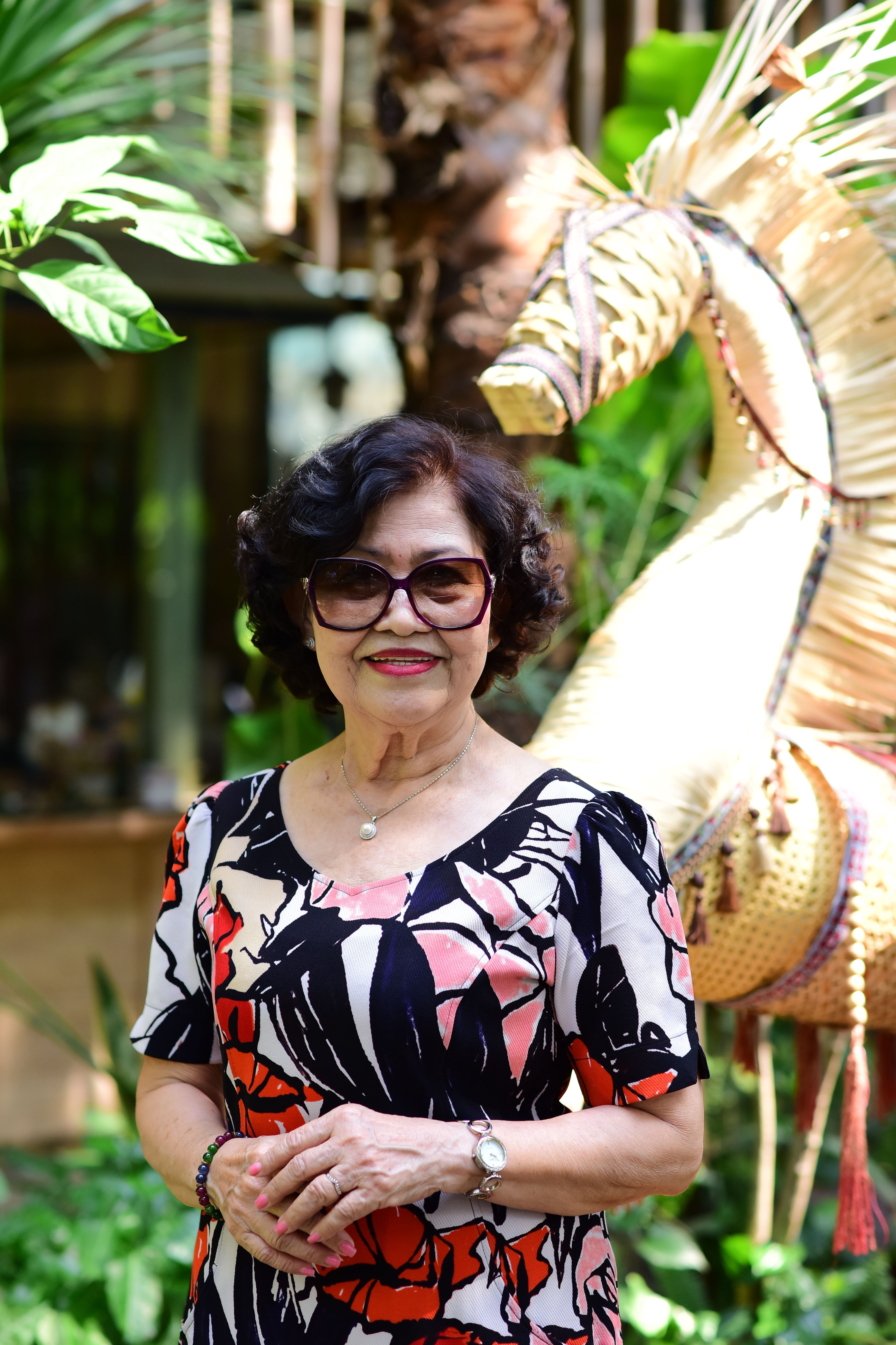A senior woman wearing stylish glasses and a floral dress poses outdoors against lush greenery.