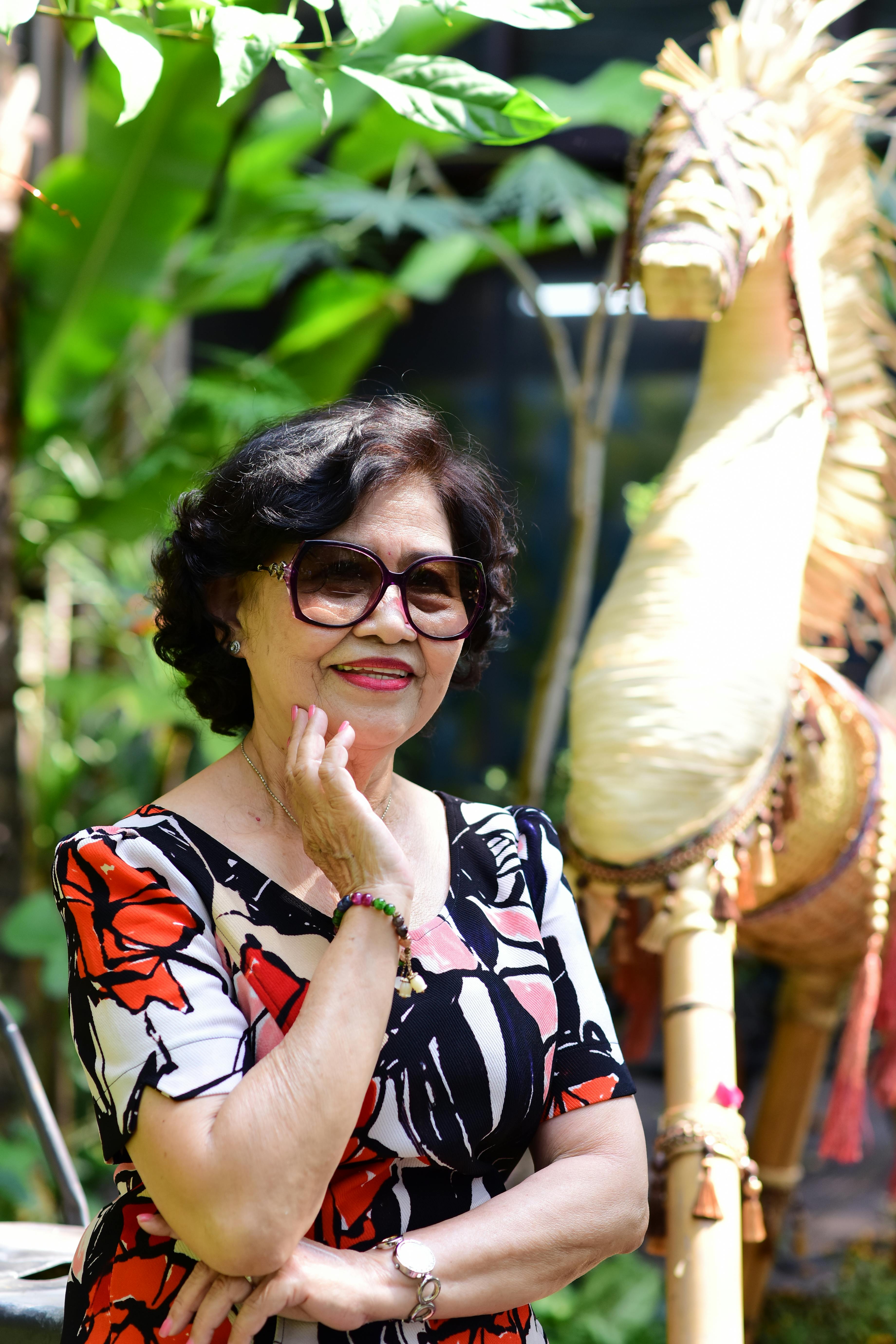 Senior woman in stylish dress poses outdoors with decorative straw horse and lush greenery.