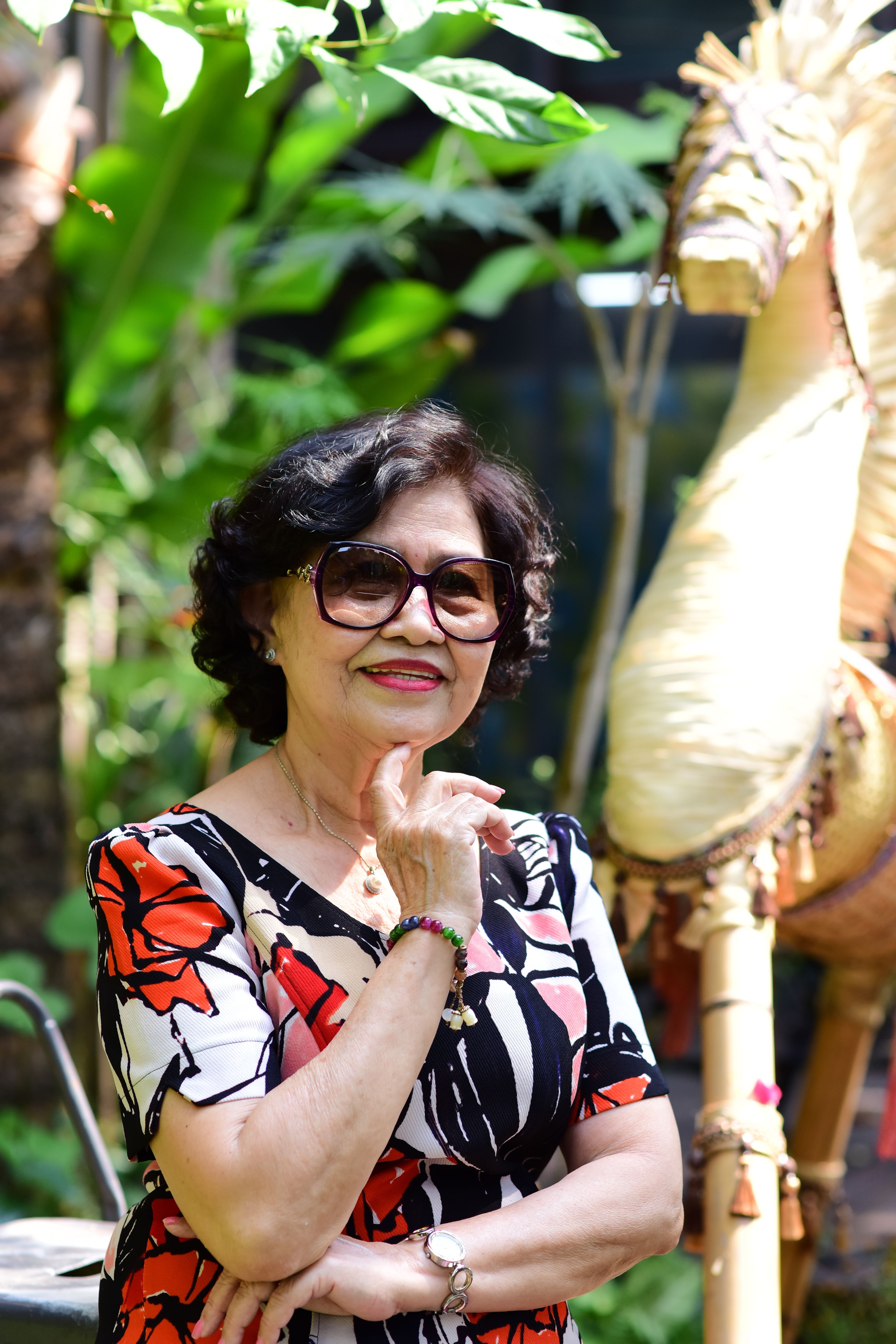 Charming senior woman in sunglasses enjoys a sunny day in a lush garden with a decorative wooden horse.