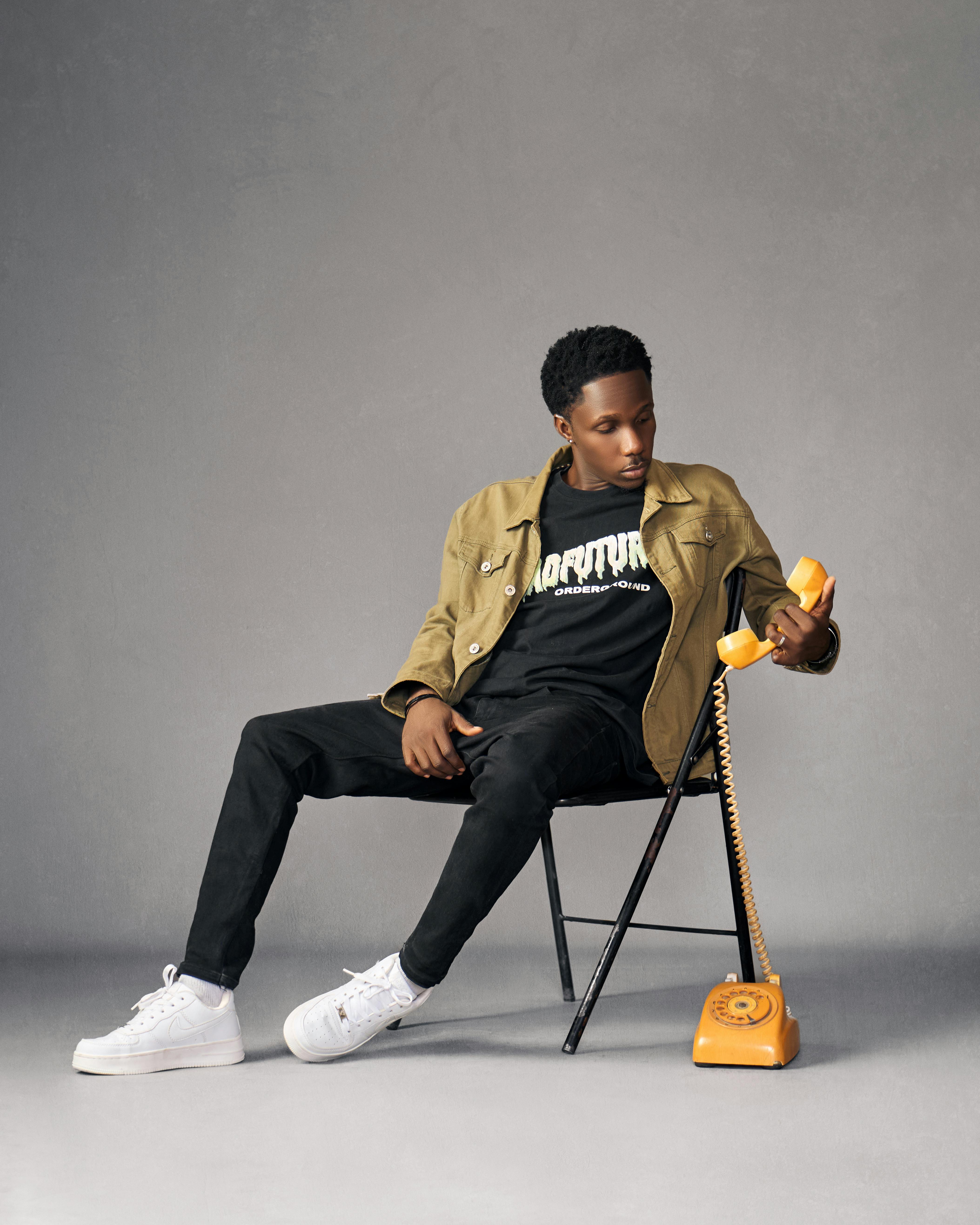 免费 一位时尚的年轻男子坐在复古橙色电话旁，展现出现代与复古的风格元素。 素材图片