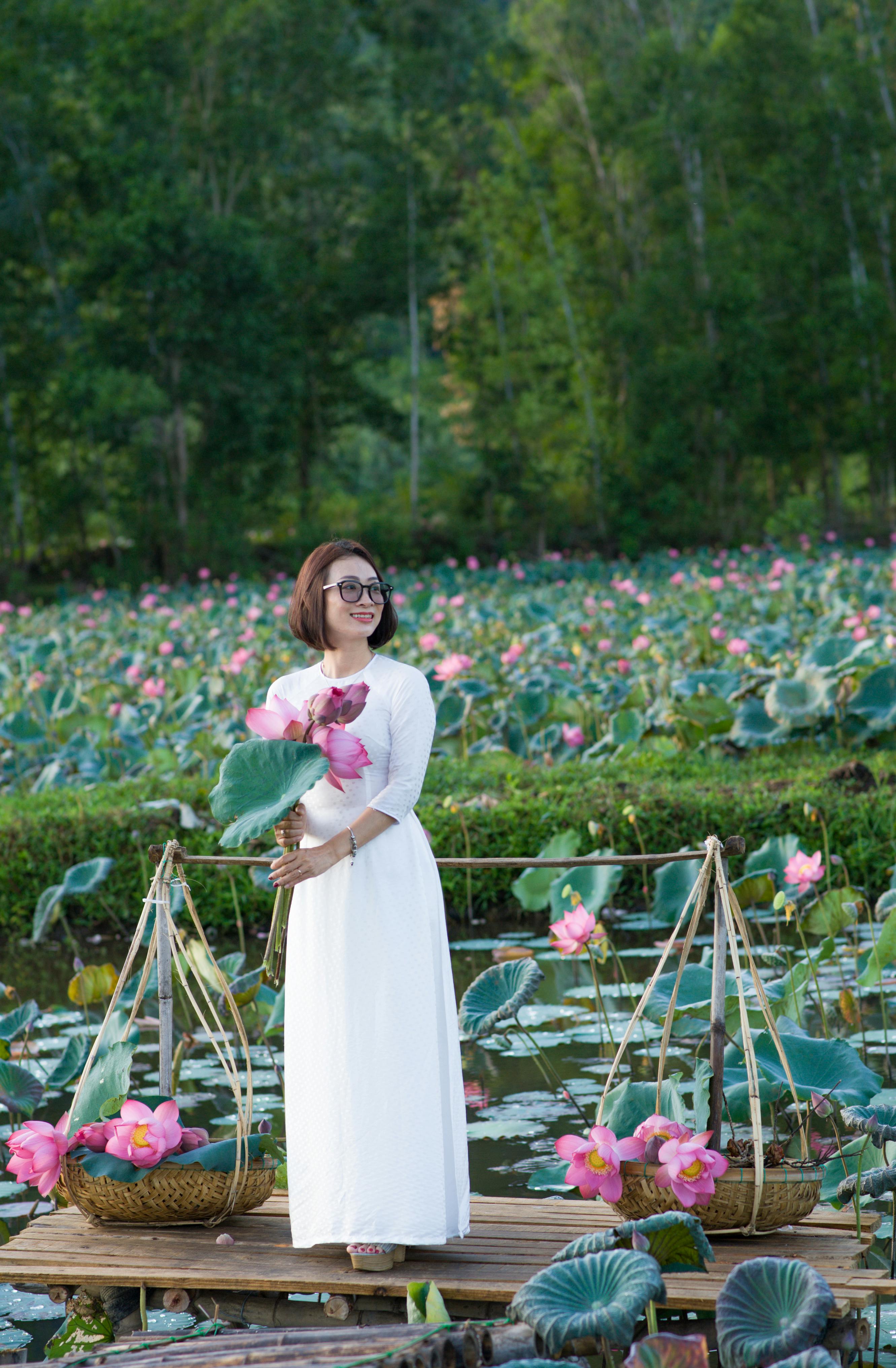 Gratis Seorang wanita berbaju putih memegang bunga teratai berdiri di tengah kolam yang rimbun. Foto Stok