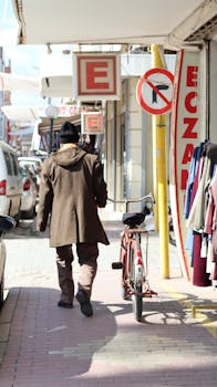 A man in a coat walks beside a bicycle down a city street lined with shops.