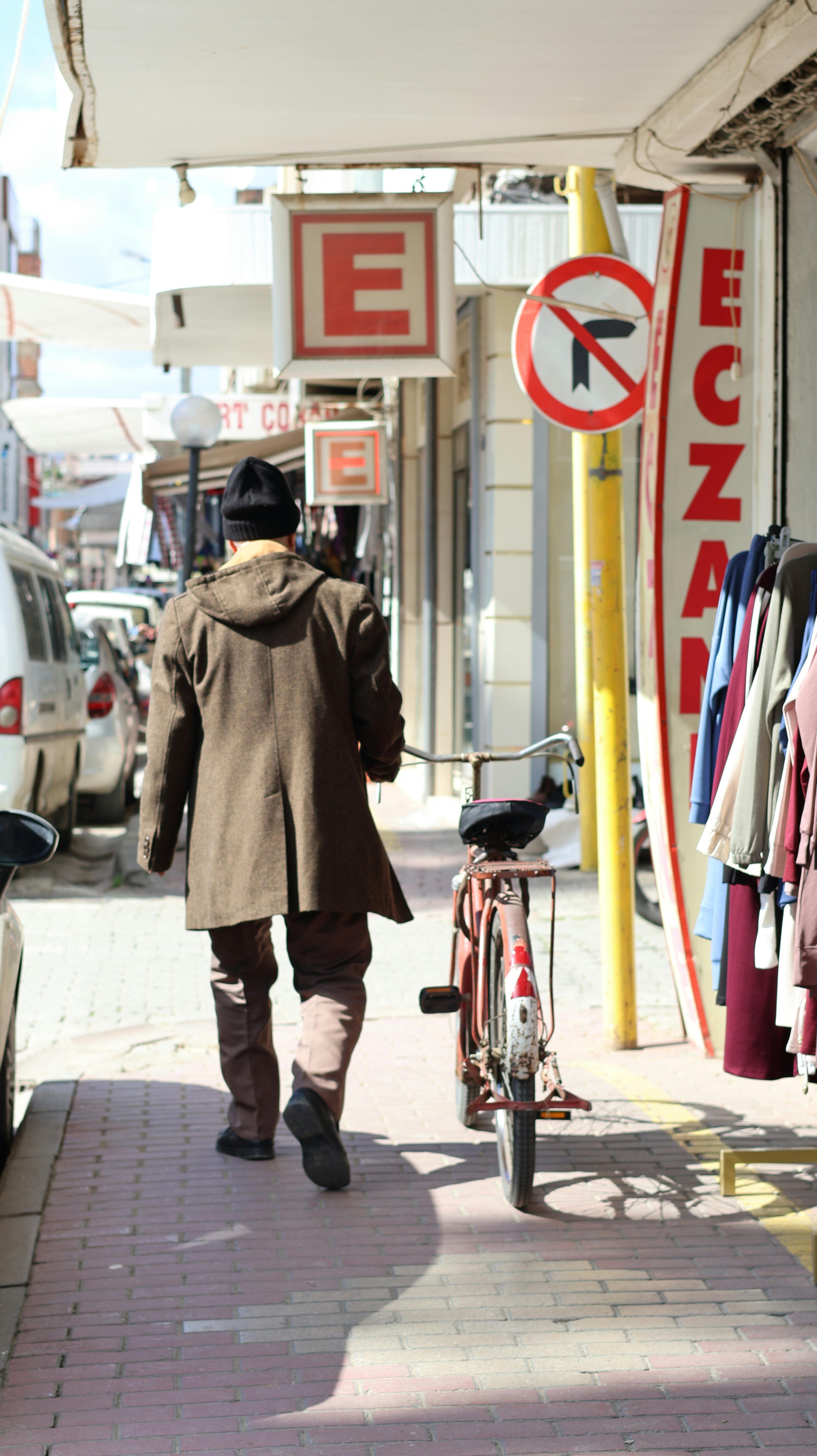 Free A man in a coat walks beside a bicycle down a city street lined with shops. Stock Photo