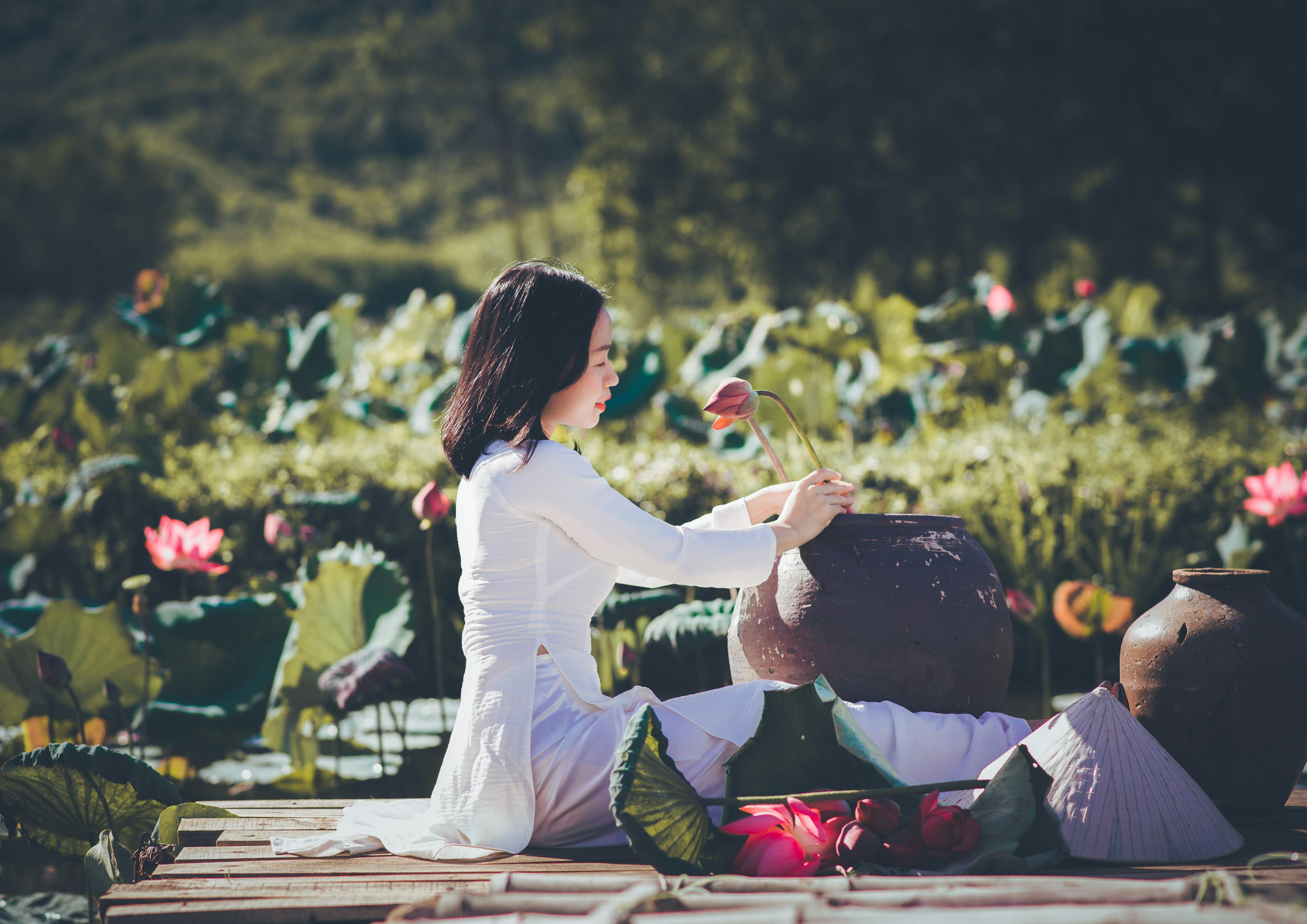Free A serene woman in traditional attire tends to lotus flowers by a pond. Stock Photo