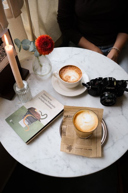 Free A cozy coffee table scene featuring cappuccino, camera, and a book on a marble table. Stock Photo