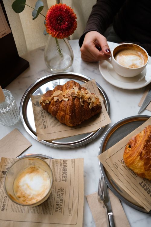 Free A warm café setting featuring almond croissants and cappuccinos on a marble table with a red flower centerpiece. Stock Photo