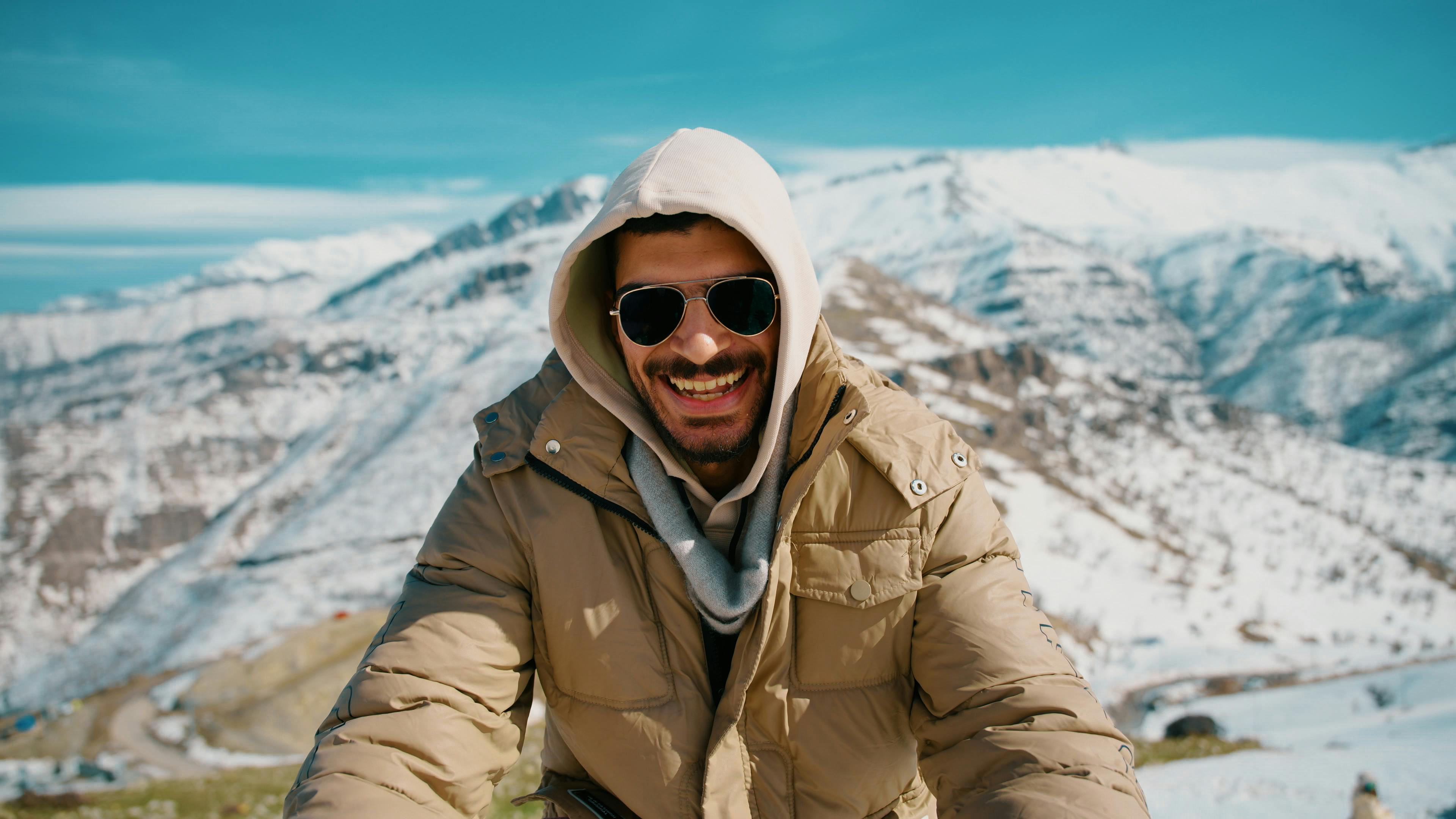 Ücretsiz Kışlık kıyafetler giymiş, gülümseyen bir adam güneşli ve karlı dağ manzarasının tadını çıkarıyor. Stok Fotoğraflar