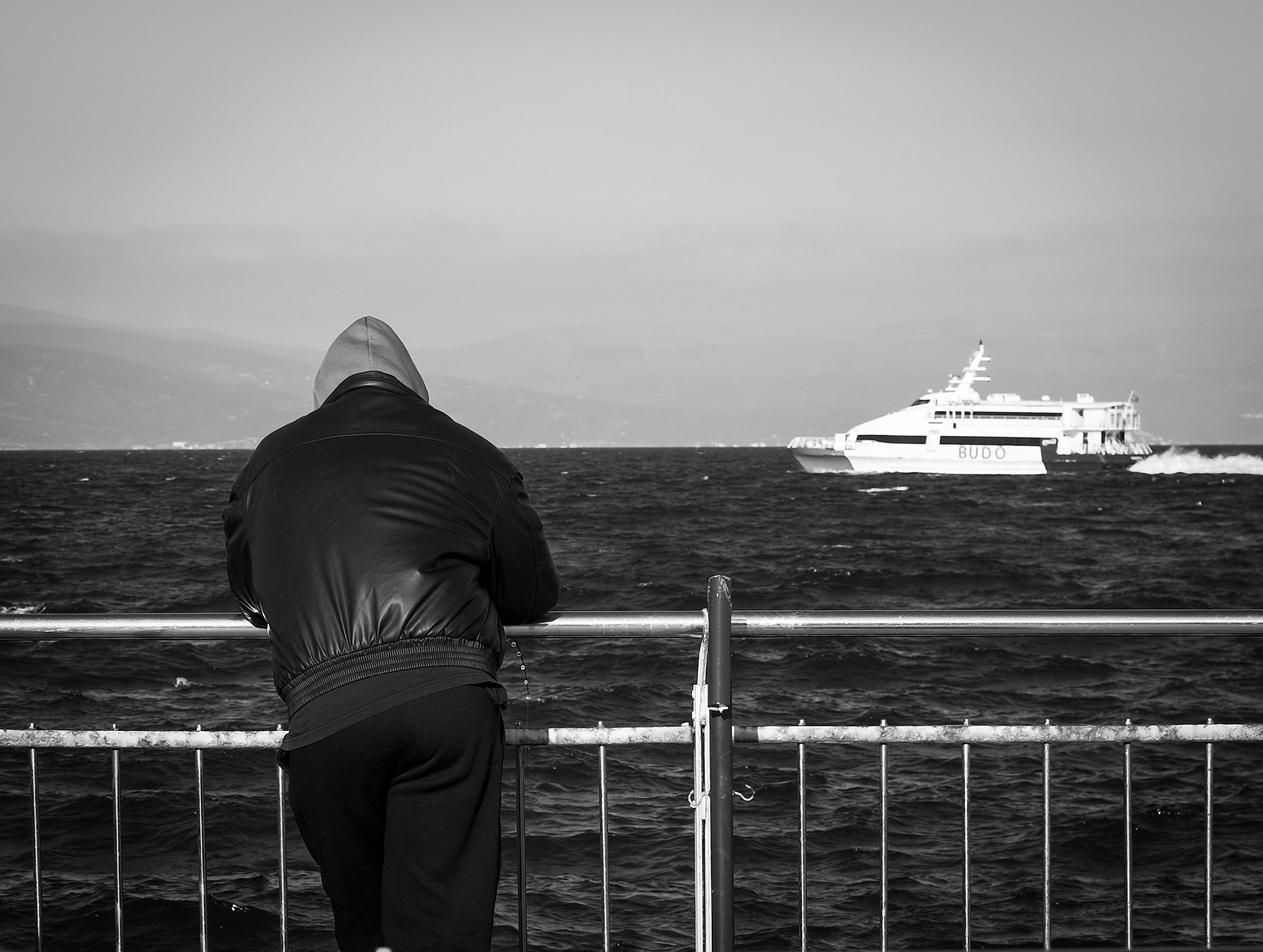 A person in a hoodie leans on a railing observing a ferry on a calm waterfront.