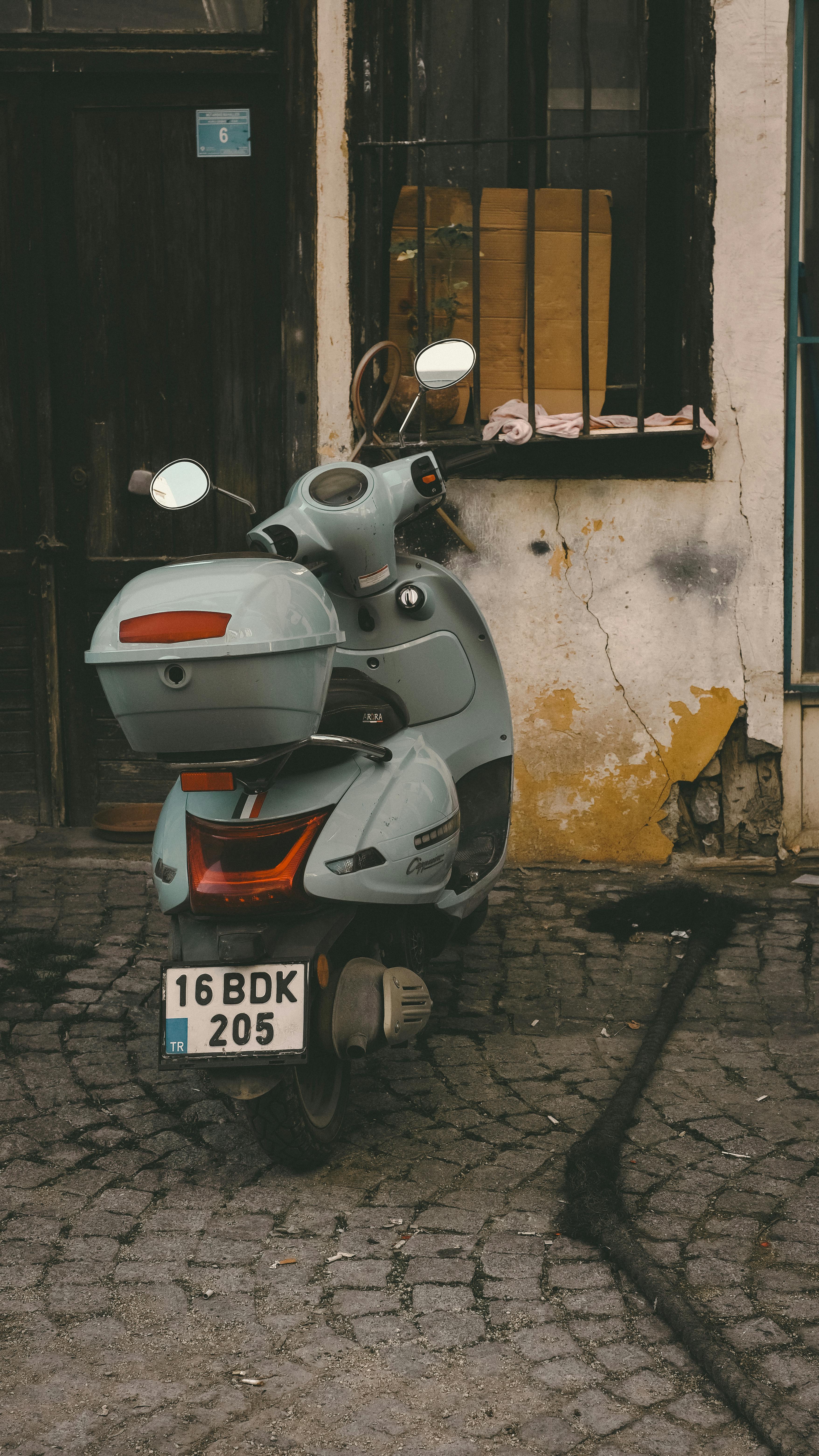 Gratis Uno scooter d'epoca parcheggiato accanto a un muro rustico e consumato dal tempo, su una strada acciottolata. Foto a disposizione