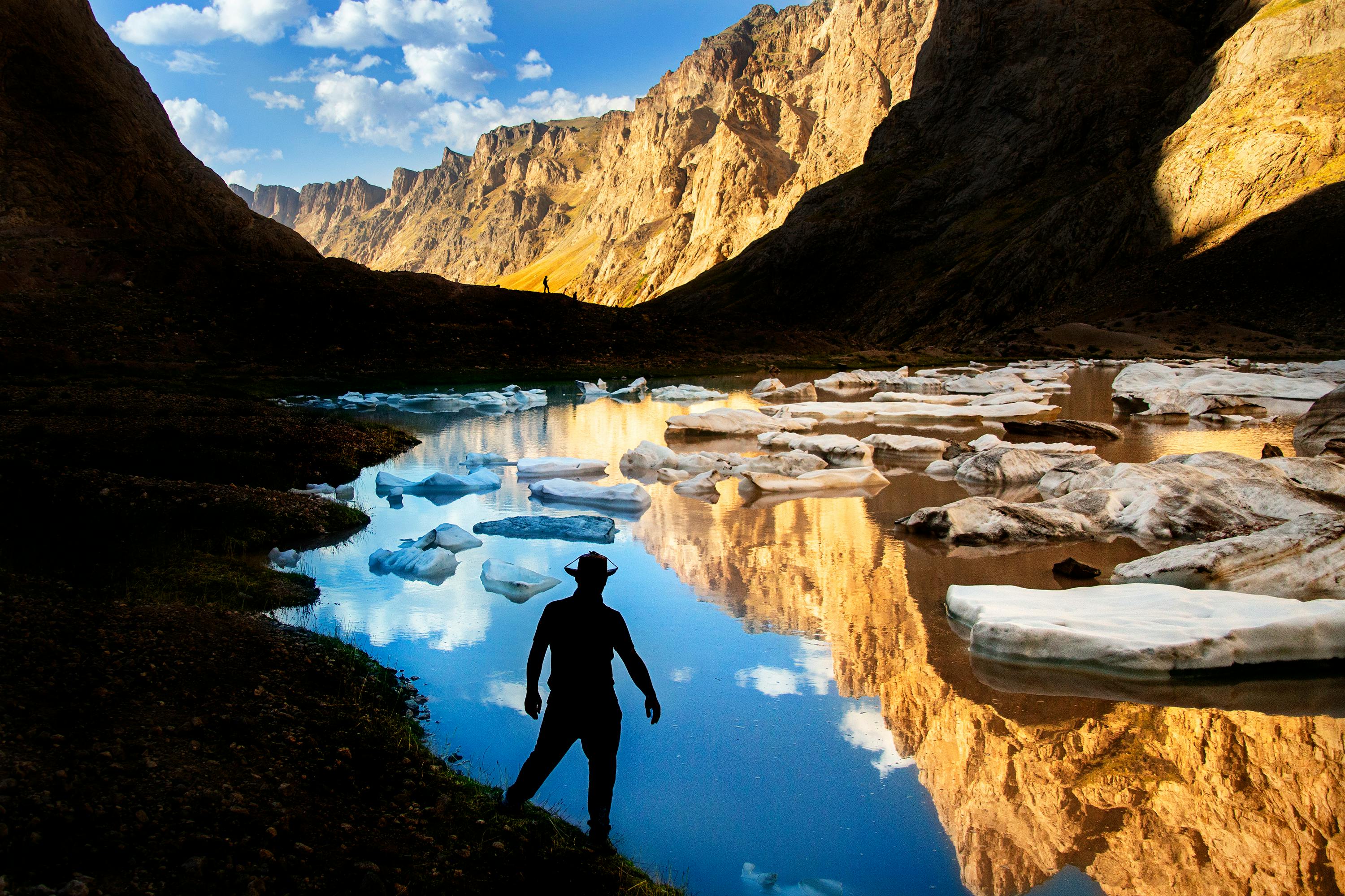 Stunning silhouette of a man by a glacier lake reflecting mountains, captured during daytime in a canyon.
