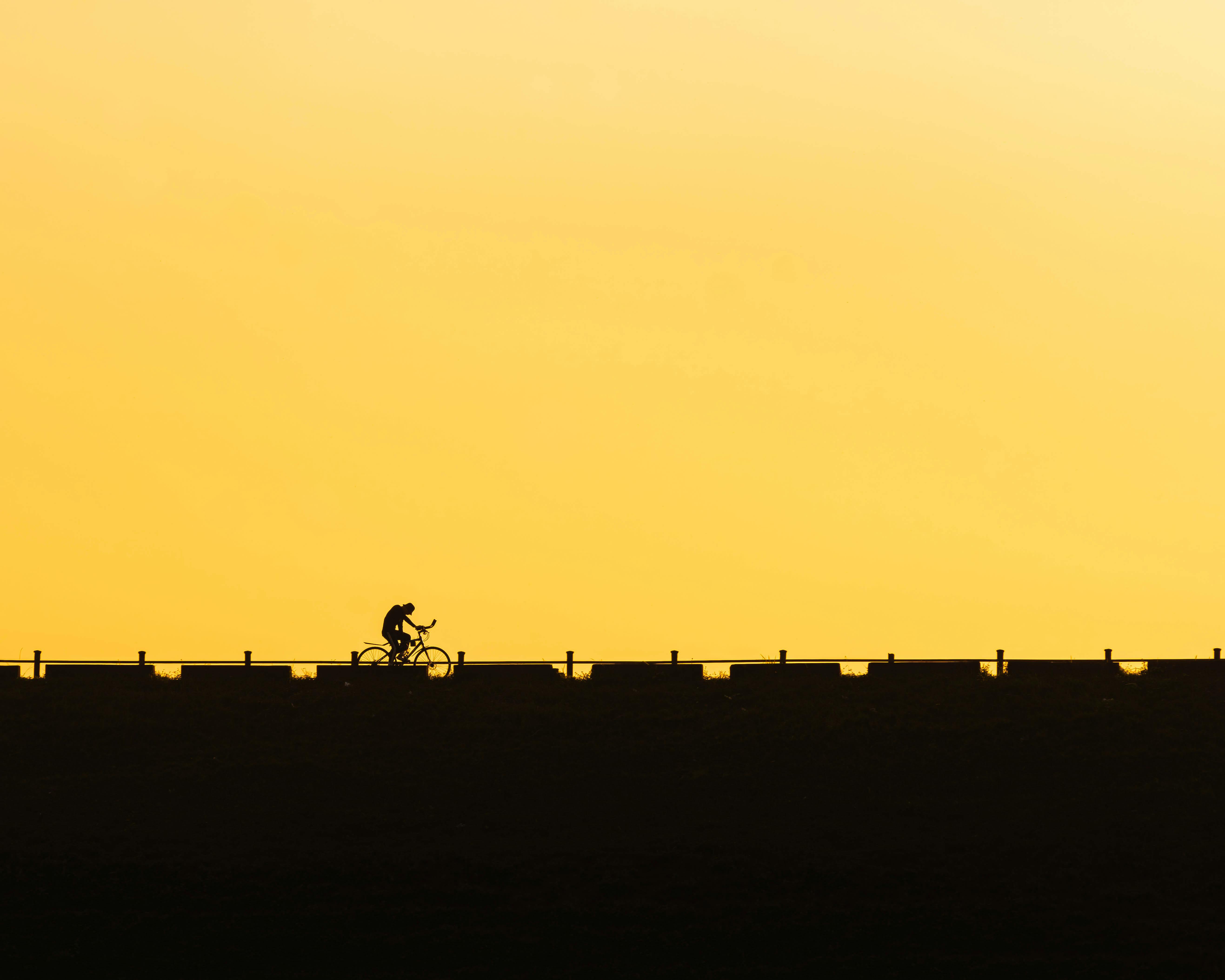 無料 鮮やかな黄色の夕焼け空を背景に、地平線に沿って走るサイクリストのシルエット。自由と冒険の感覚を捉えています。 写真素材
