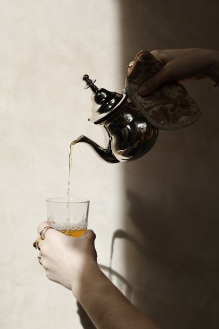 A silver teapot pouring tea into a glass, casting shadows.