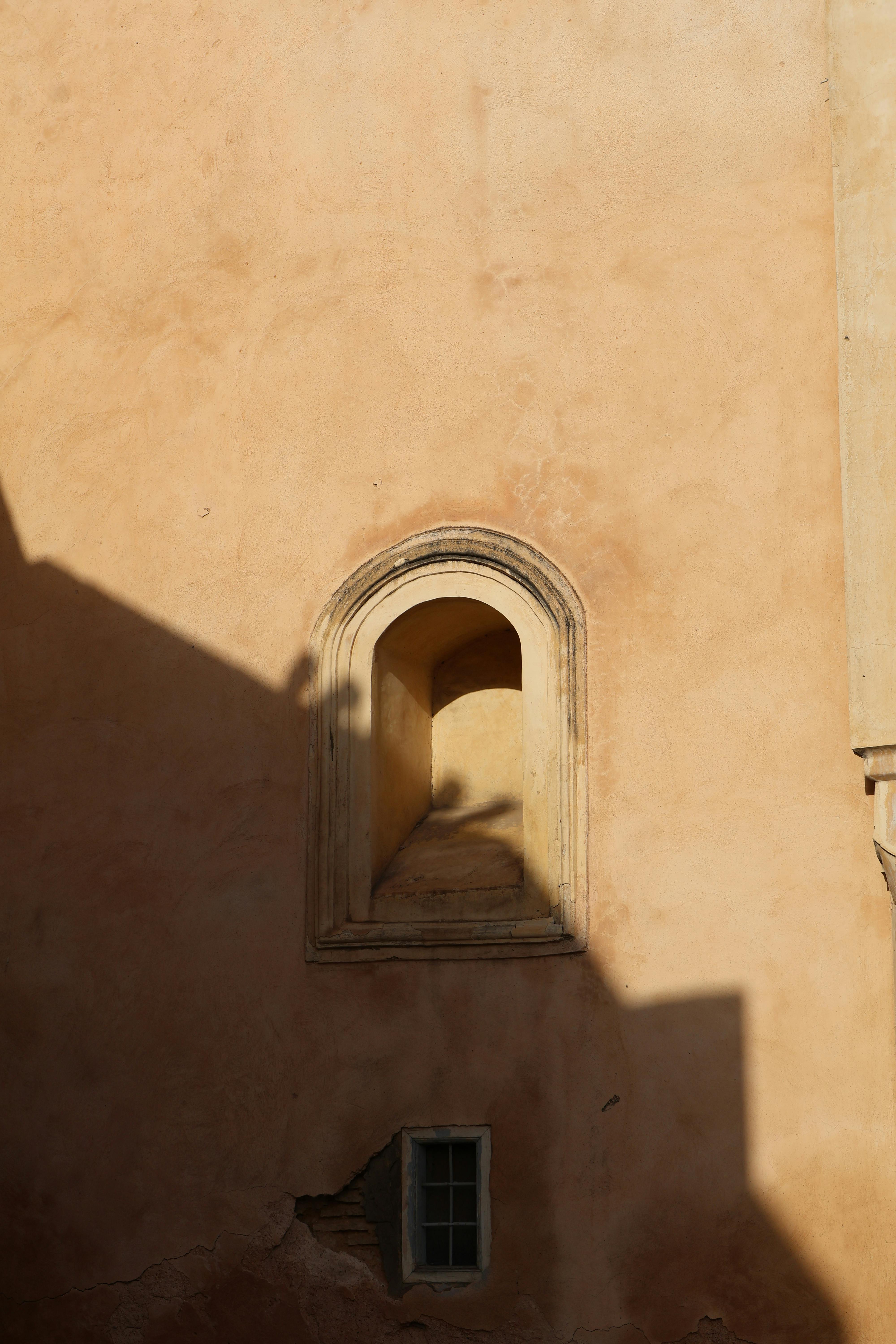 Gratis Un muro rustico con una finestra ad arco che proietta ombre intense sotto la calda luce del sole. Foto a disposizione