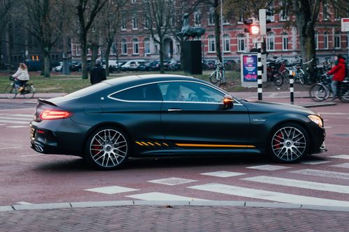 Free A sleek black sports car driving in a vibrant city setting with people and bicycles around. Stock Photo